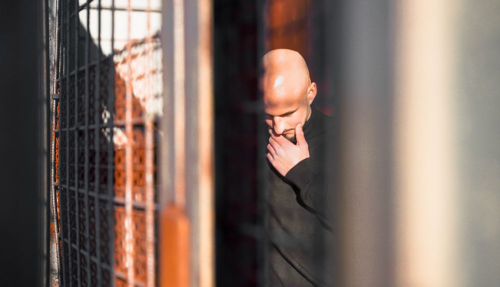 Bald man in black outfit seen through metal fence bars in dramatic urban environment.