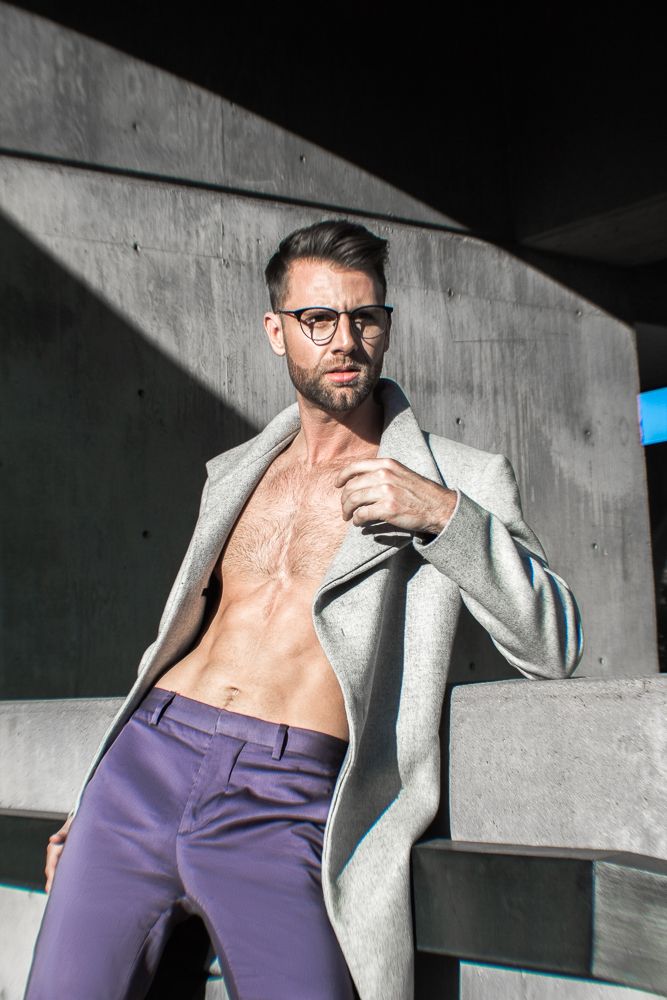 Bearded man in glasses wearing open grey coat with bare chest leans against concrete wall.