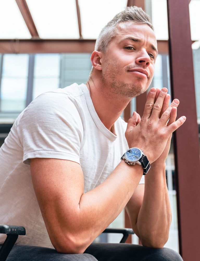 Athletic blonde man in white t-shirt wearing a blue watch with hands clasped together.