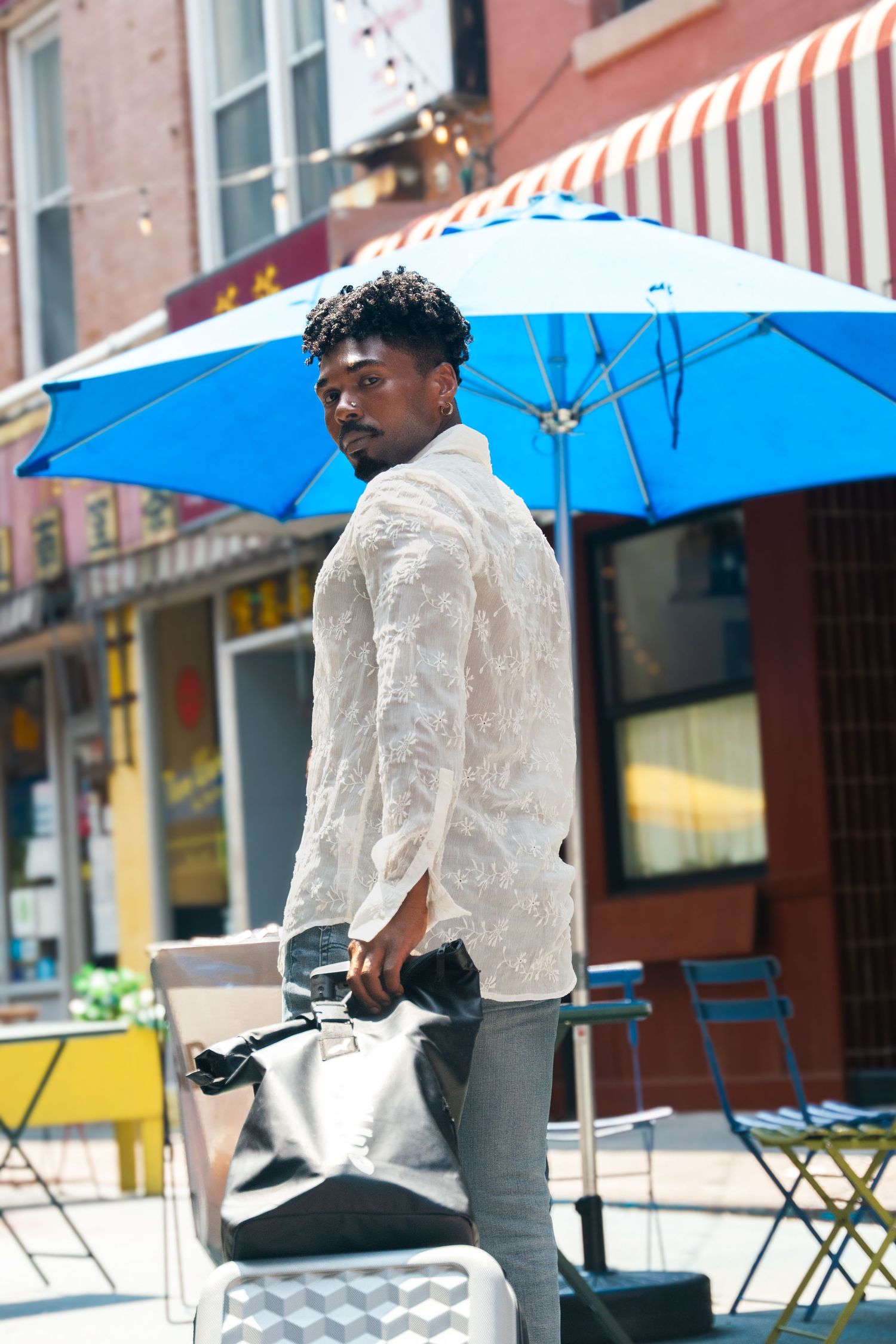 Stylish man in white lace jacket holding a bag on a sunny urban street with a blue patio umbrella.