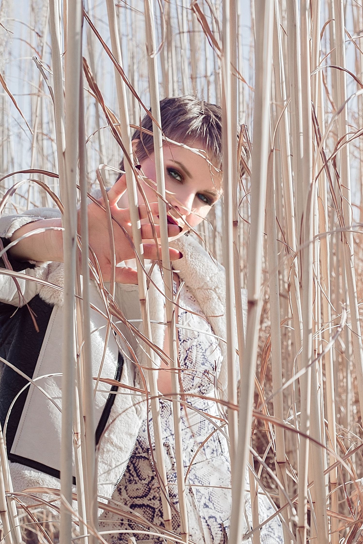 Elegant woman in a lace dress peering through tall dry reeds in a soft natural light setting.