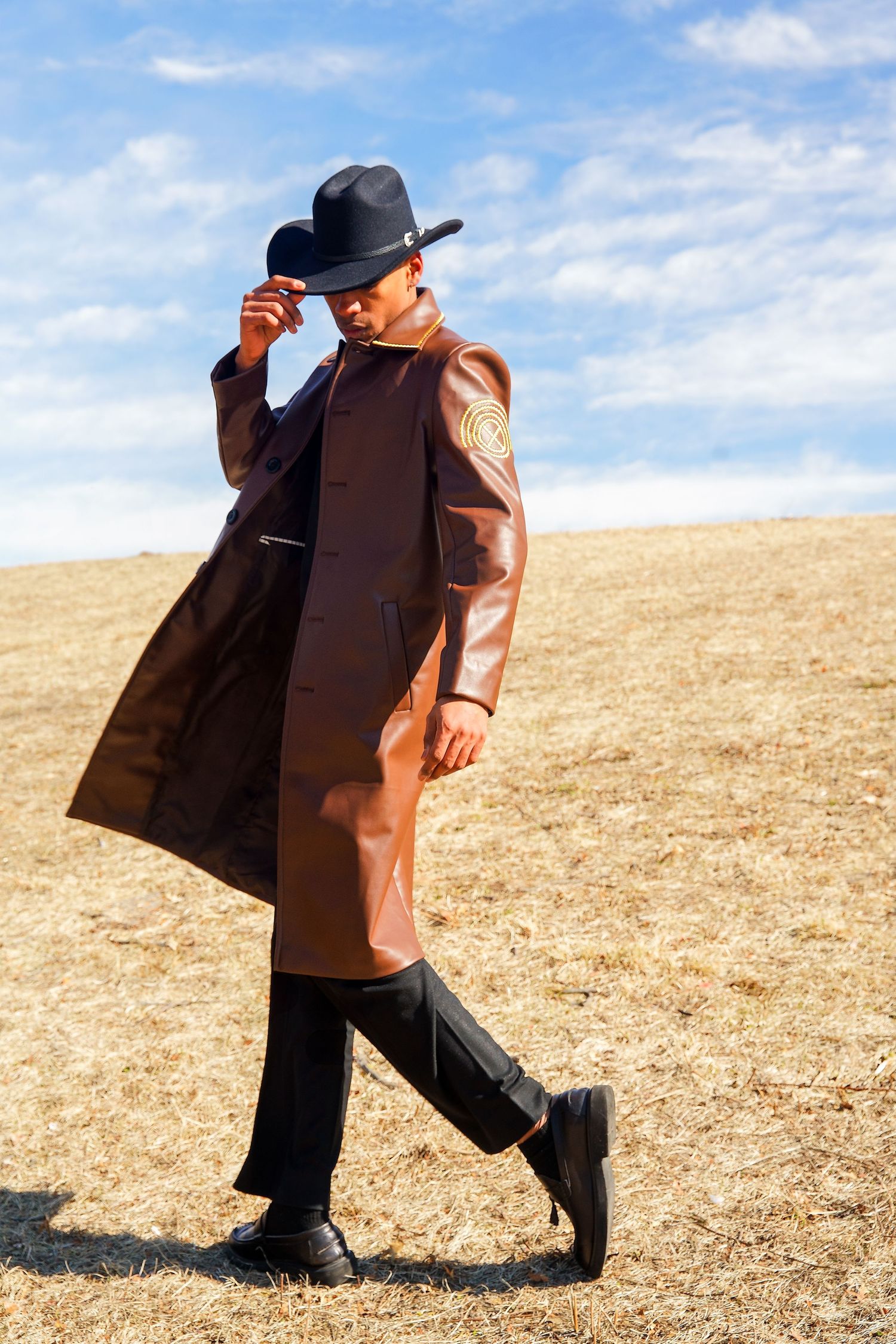 Stylish man in brown leather duster coat and black cowboy hat walks across dry field under blue sky.