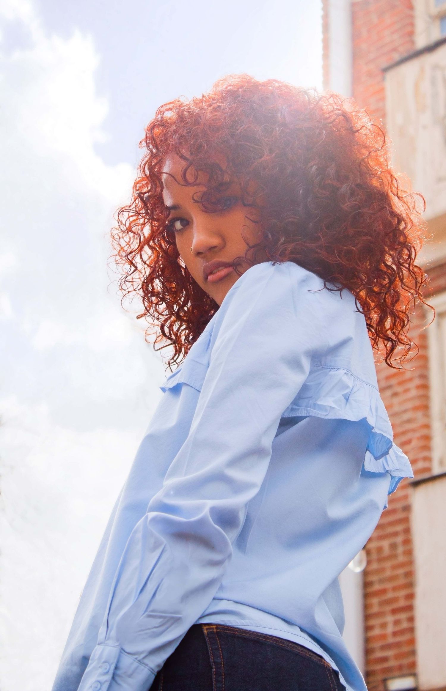 Young woman with vibrant red curly hair wearing a light blue ruffled shirt, posing outdoors near a brick building.