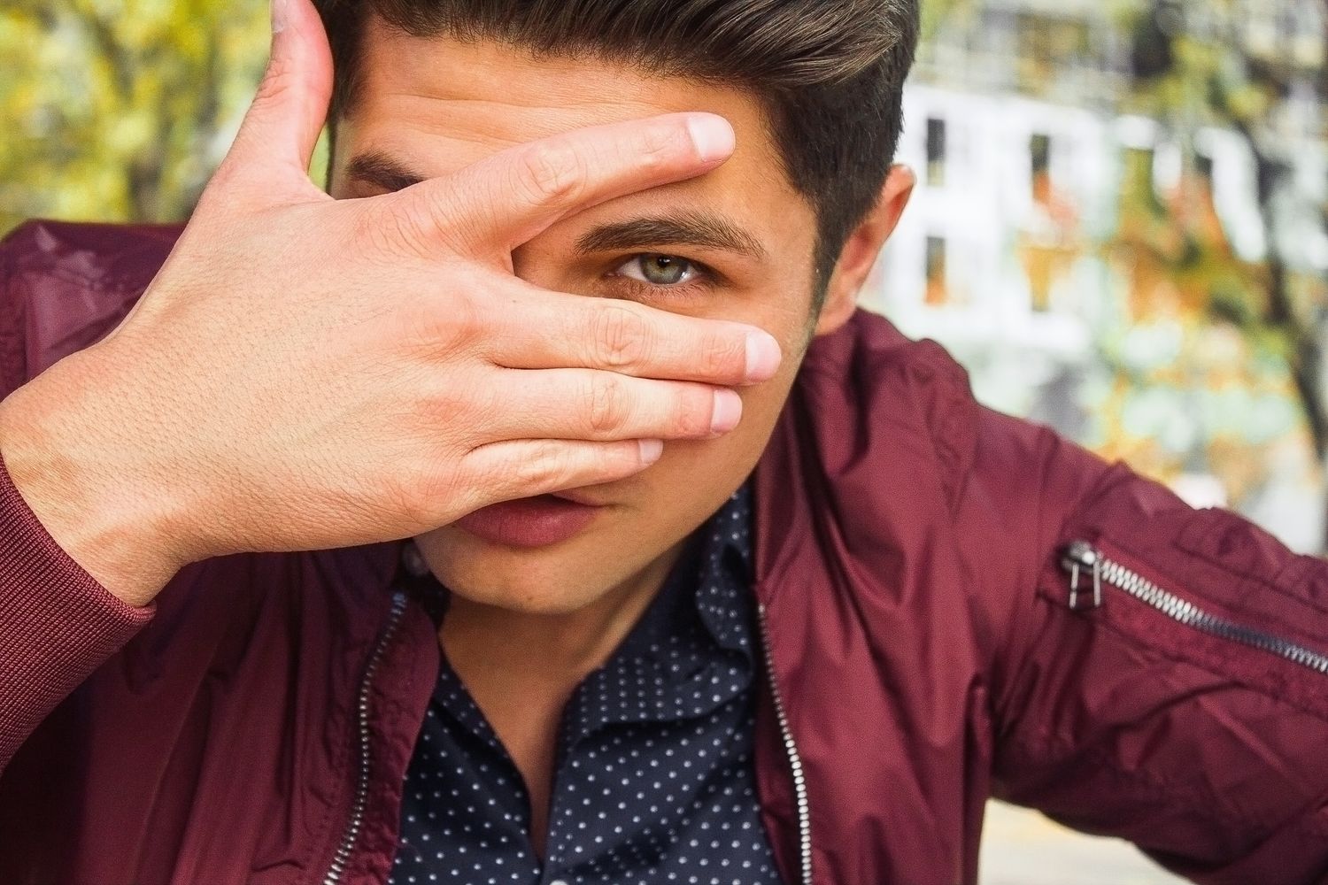 Young man in red jacket peeking through fingers covering his face outdoors.