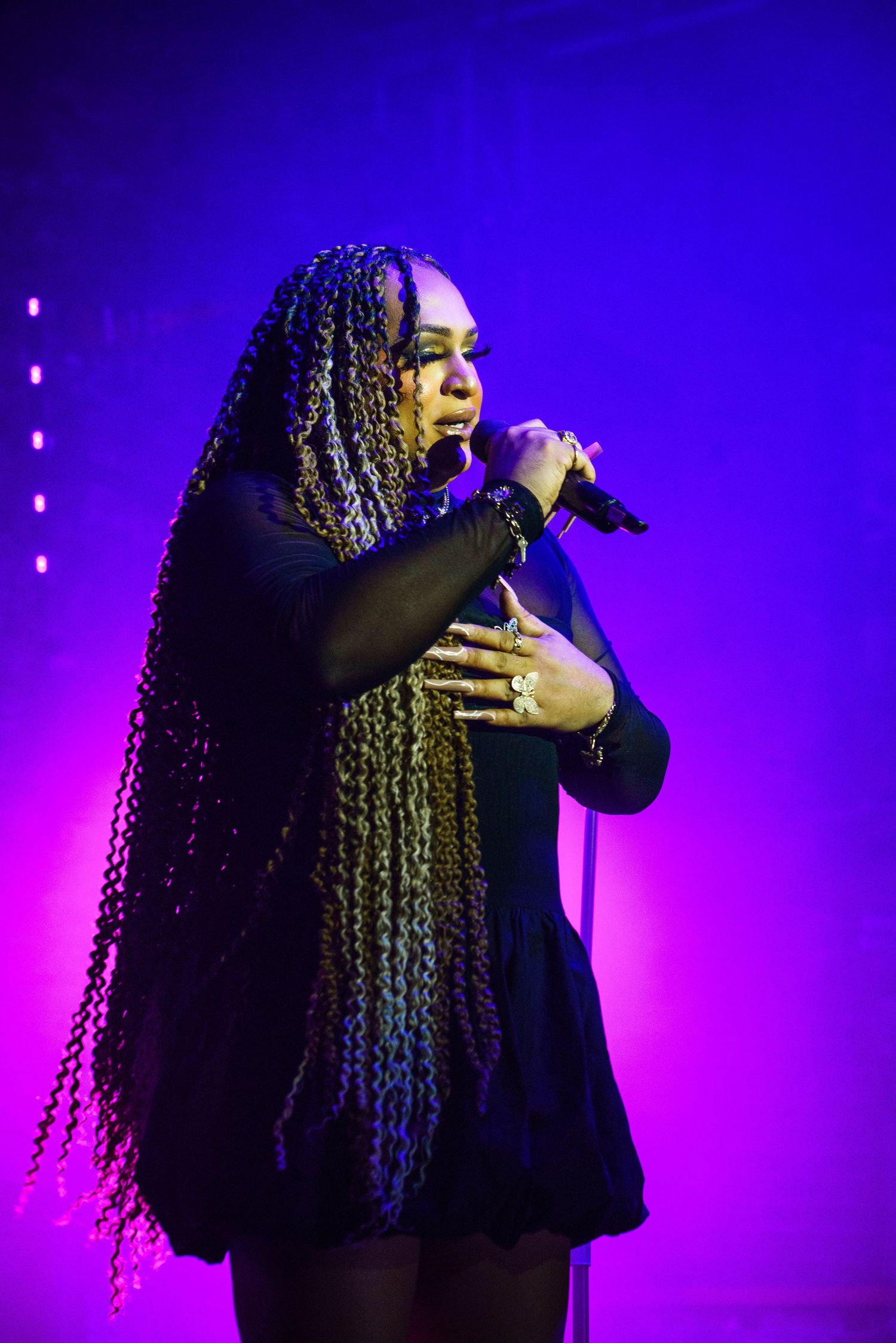 Female singer with long braided hair performs on stage holding microphone, illuminated by purple and pink stage lights.