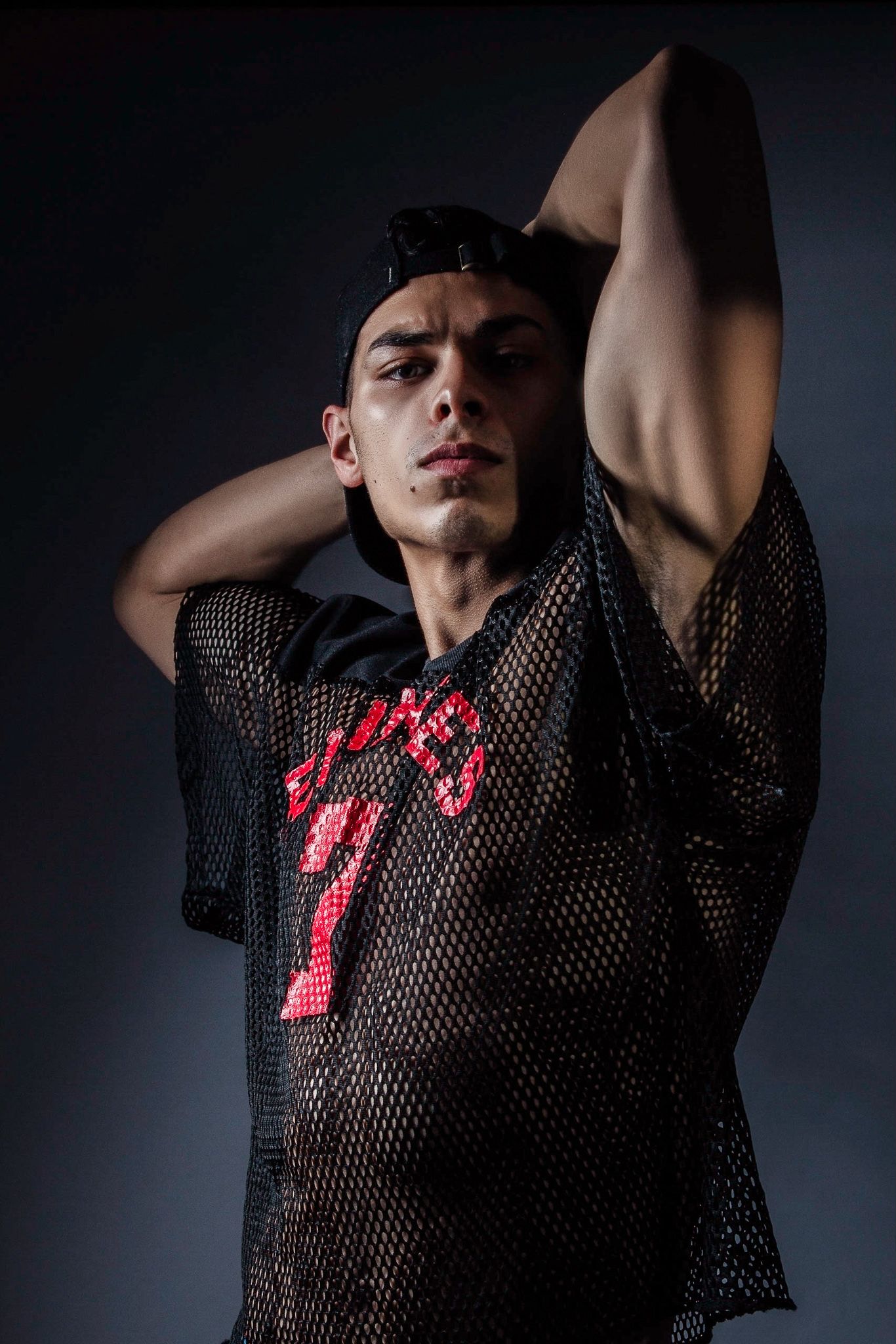 Young man in black mesh jersey with red number, hands behind head, posing against dark studio background.