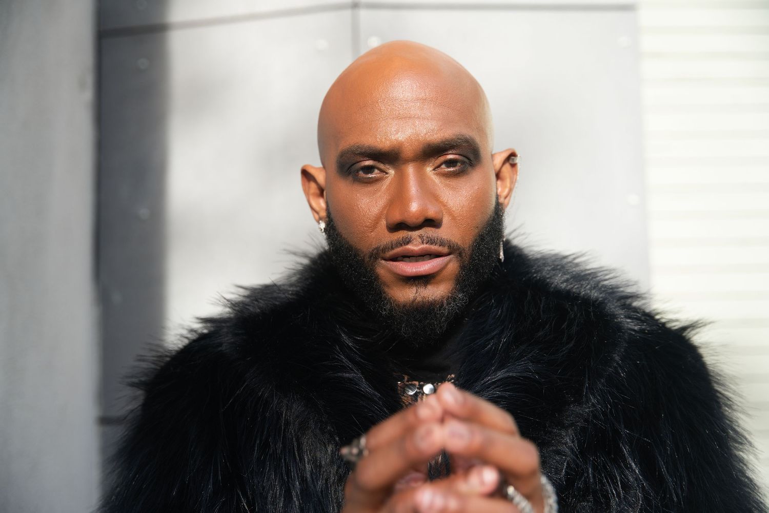 Bald man with beard wearing black fur coat, hands clasped, posing confidently against a white wall.
