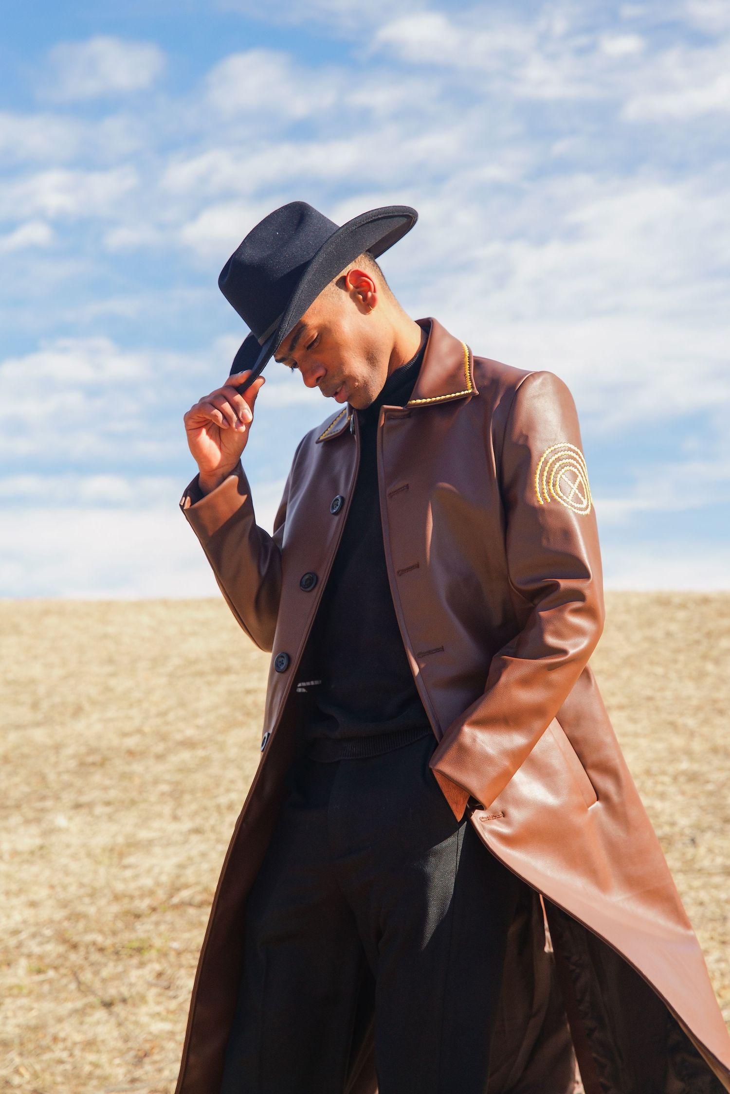 Stylish man in brown leather trench coat and black hat posing in an open field under blue sky.