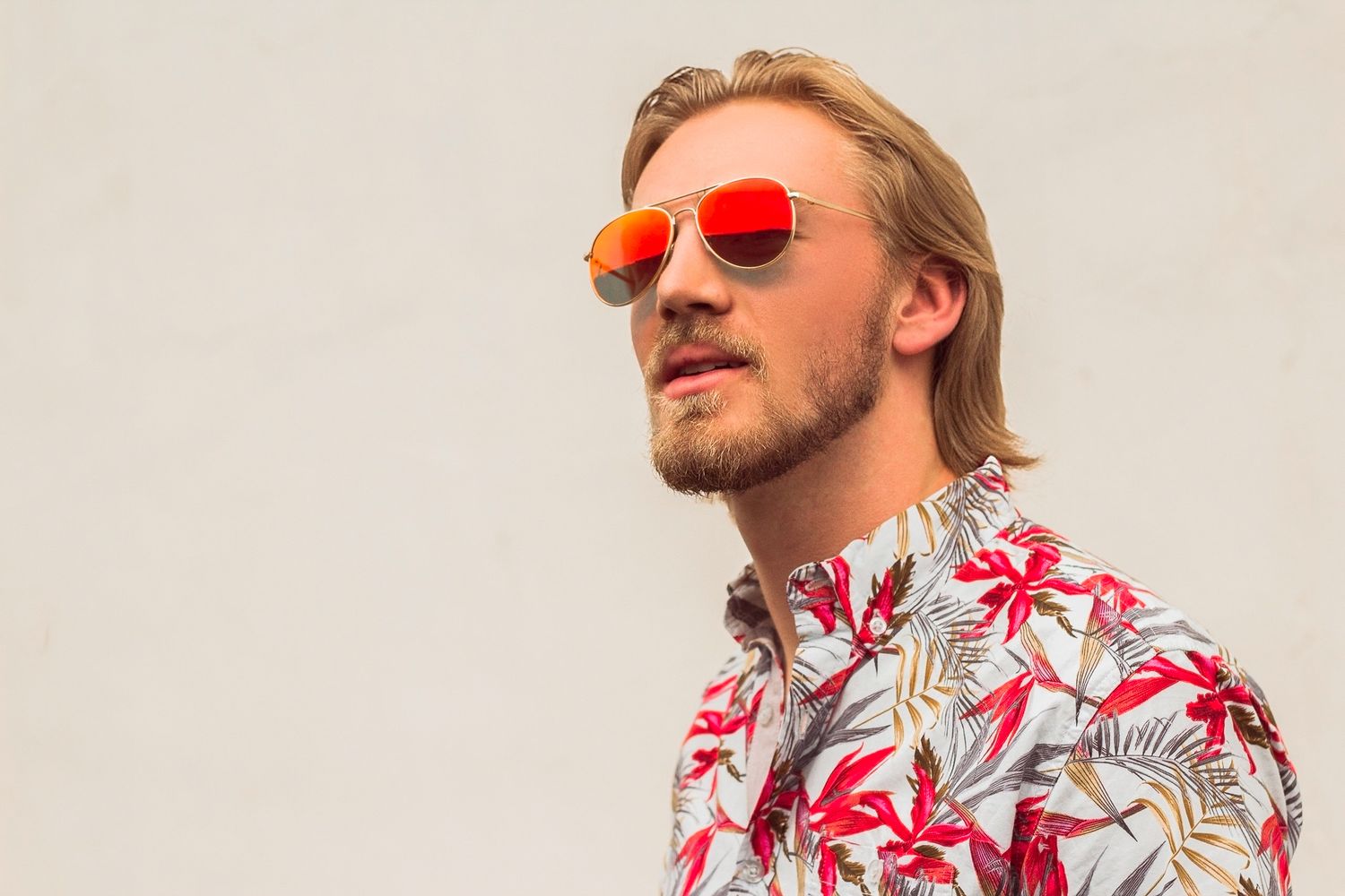 Blonde man with beard wearing red floral shirt and mirrored aviator sunglasses against white wall.