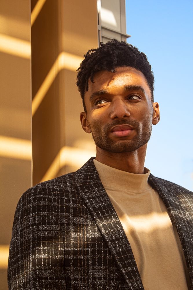 Young Black man in plaid blazer stands by golden wall in warm sunlight looking upward.