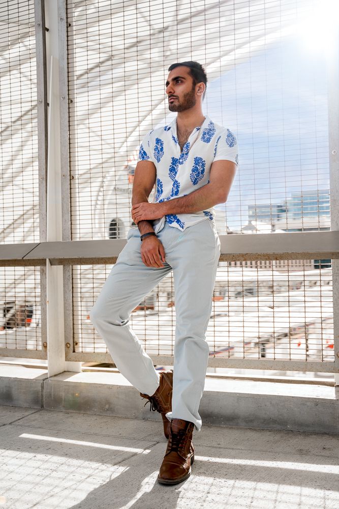 Fashionable man in blue floral shirt and white pants leaning on railing in modern urban setting.