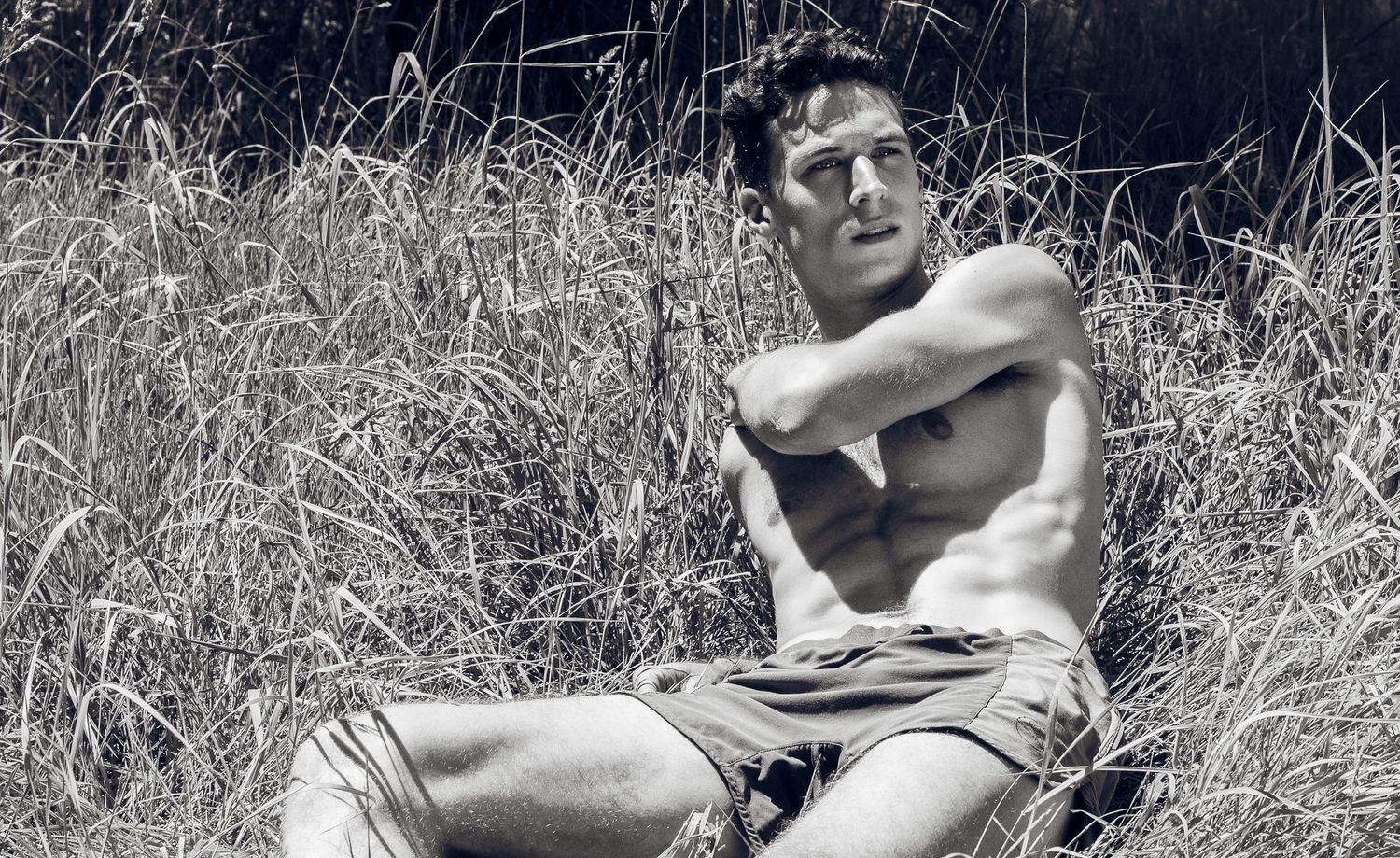 Black and white photo of a shirtless muscular man sitting in tall dry grass outdoors.