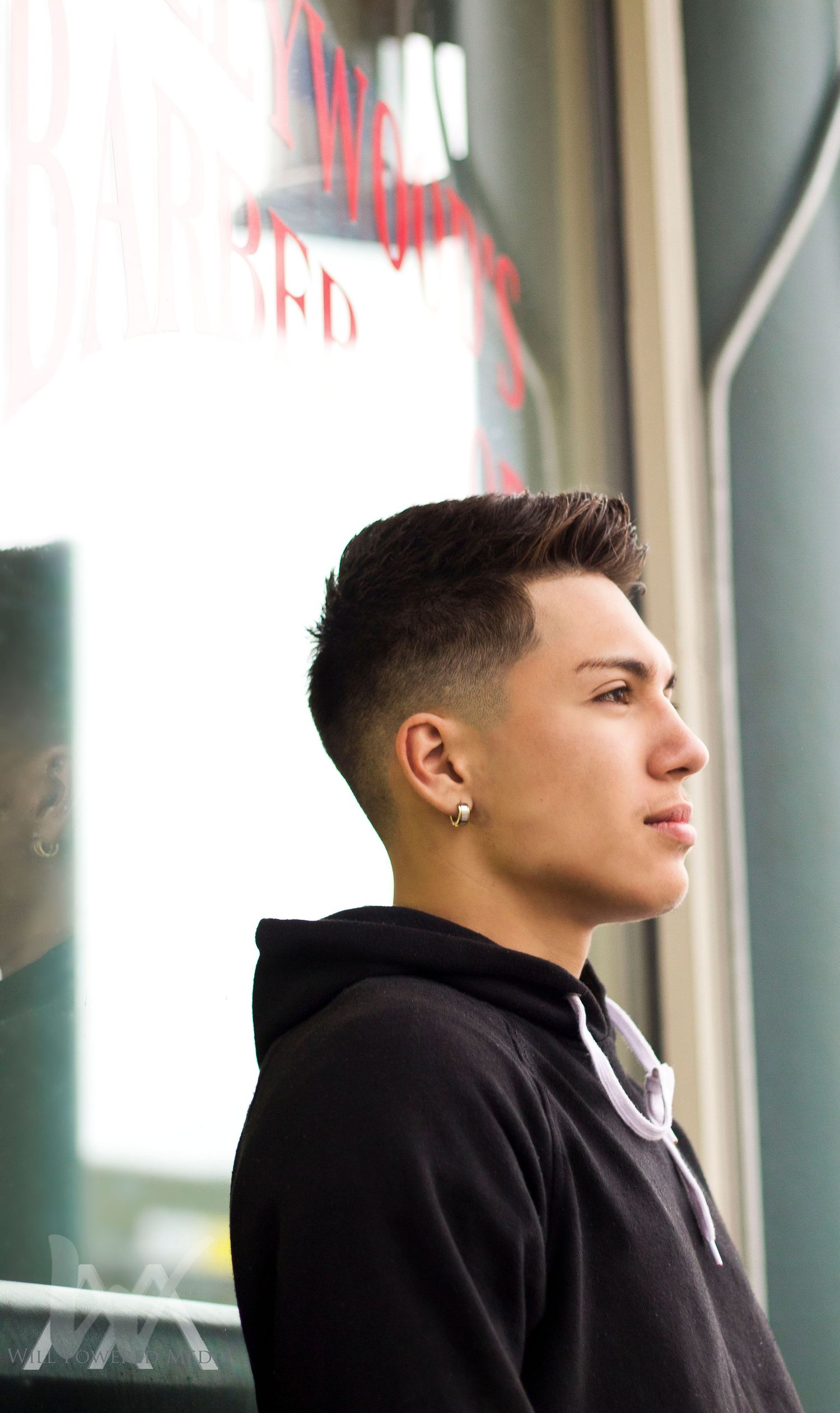 Young person with fade haircut in black hoodie gazing sideways outside a barbershop.