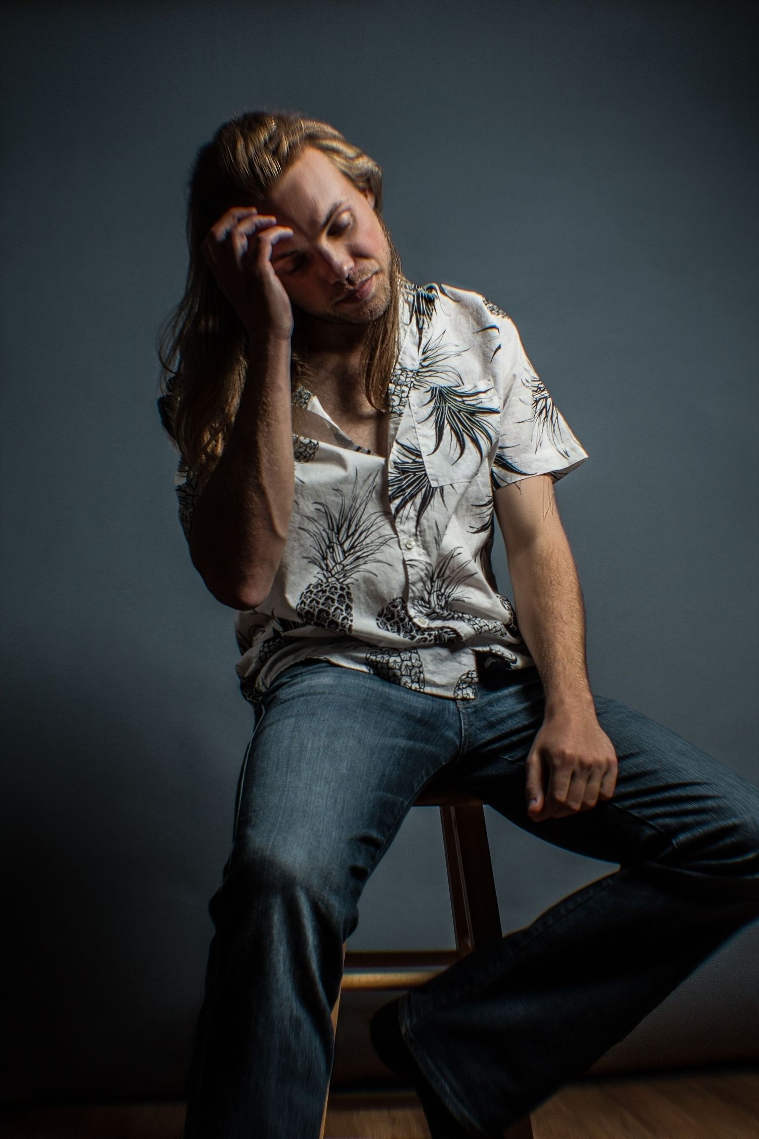 Long-haired man in pineapple print shirt sitting on stool against gray studio background.