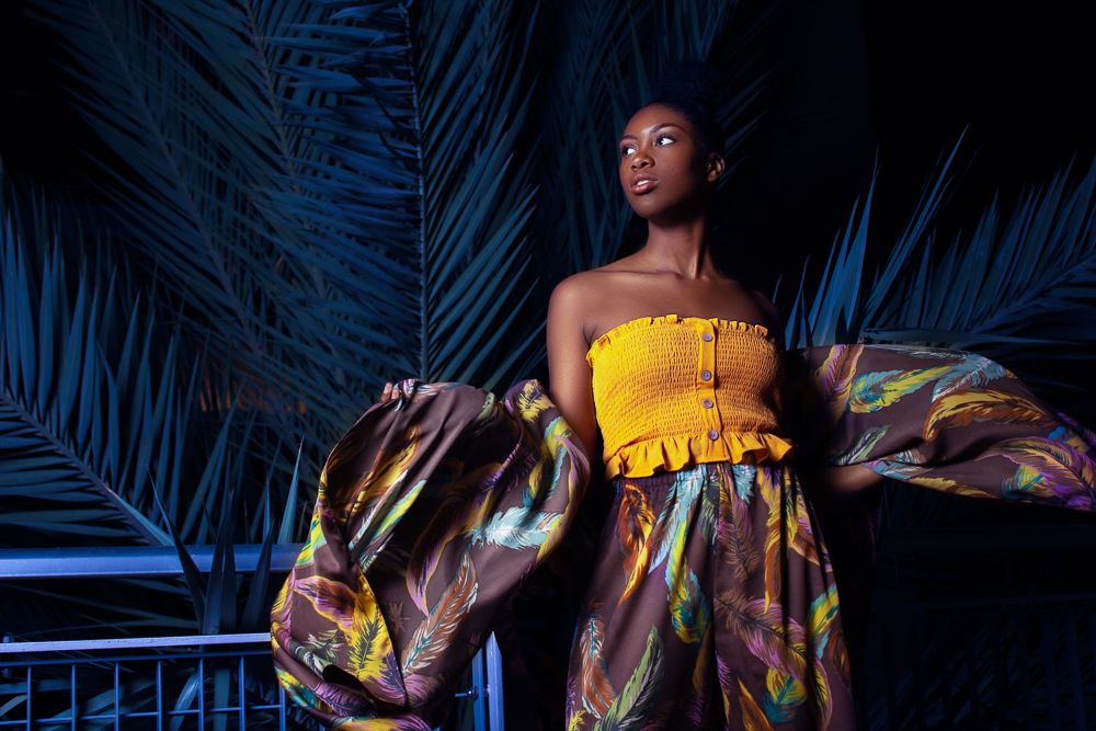 A Black woman in a yellow strapless top and colorful skirt poses among tropical blue palm leaves.