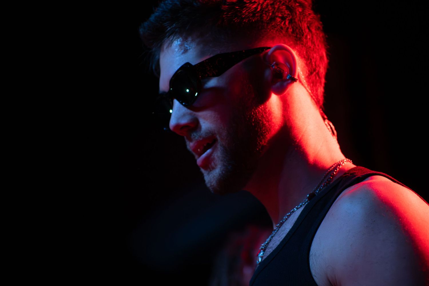 Young man wearing futuristic sunglasses and a tank top under dramatic red and blue neon lighting.