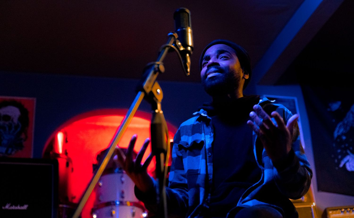 A musician recording vocals in a dimly lit studio with a condenser microphone and Marshall amp in the background.