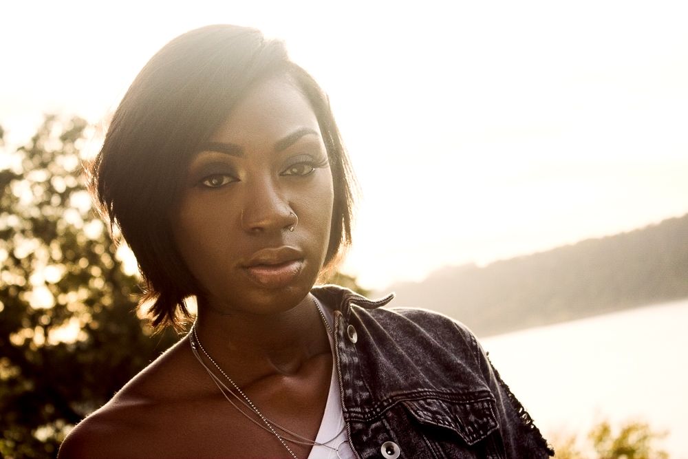 Young Black woman with bob haircut in denim jacket with golden backlight near water.