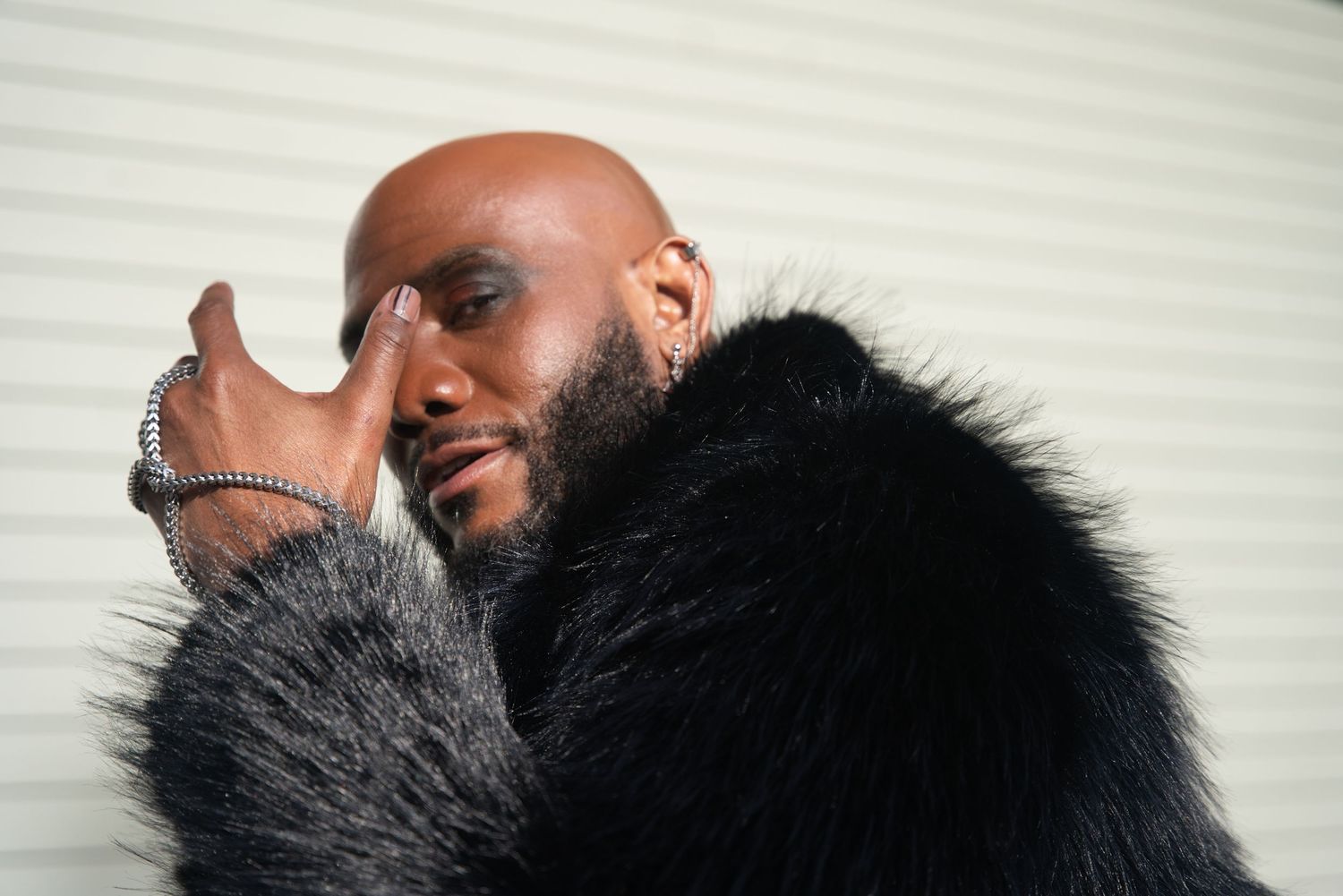 Bald man in luxurious black fur coat posing with hand near face, wearing silver rings, against white background.