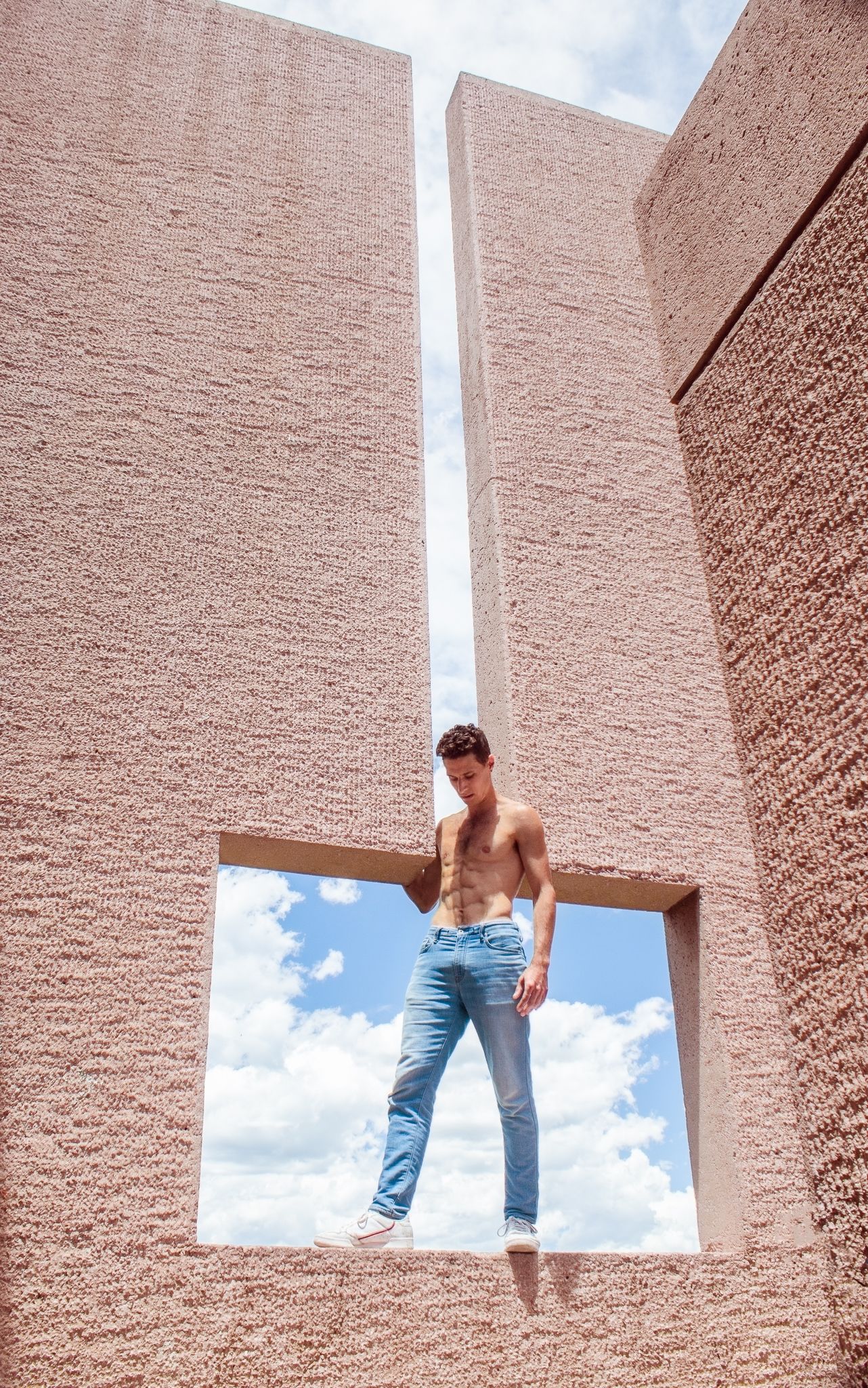 Shirtless muscular man in blue jeans posing in a square window opening of a pink textured brick wall.
