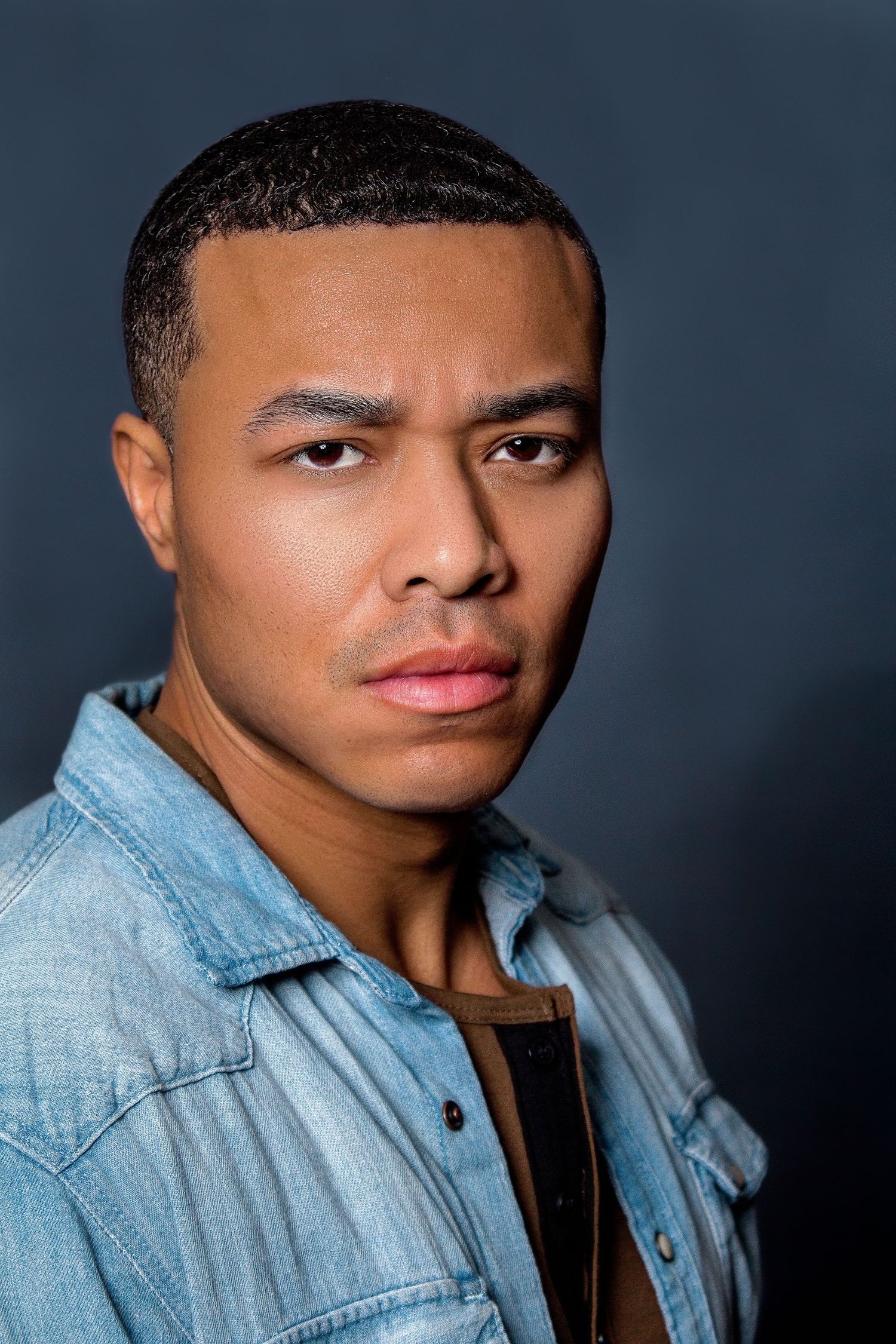 Young man in denim shirt poses against dark gray background in a serious studio portrait photo.