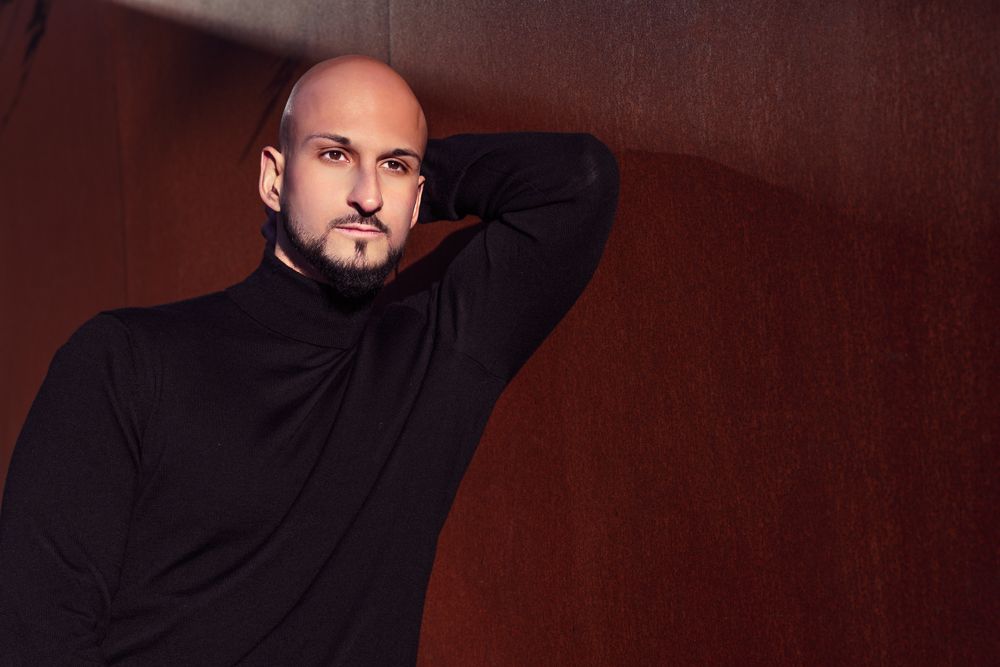 Bald man with a beard wearing a black turtleneck sweater posing against a dark wooden background.