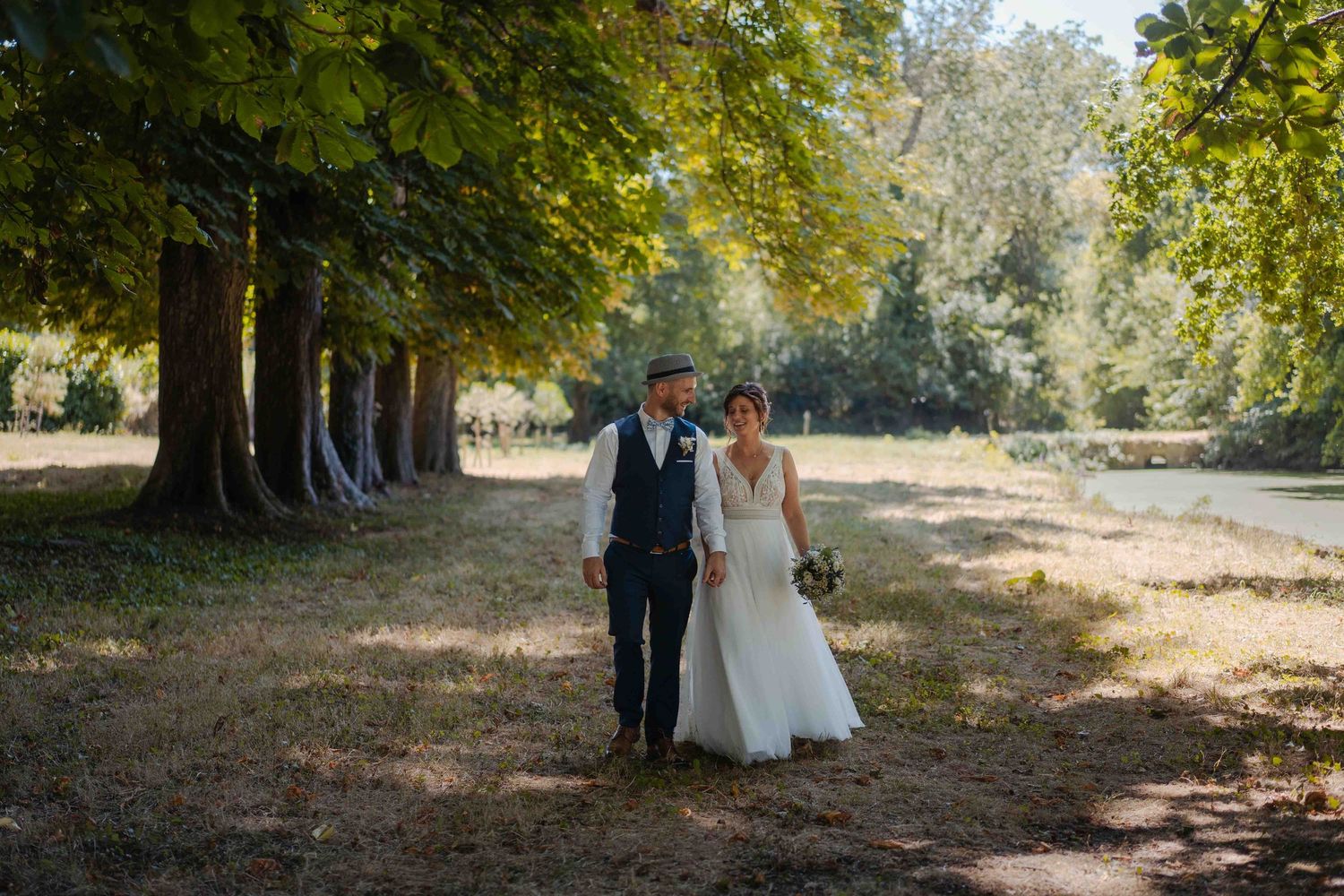 Les mariés se promènent main dans la main sous de grands arbres dans un parc ensoleillé.