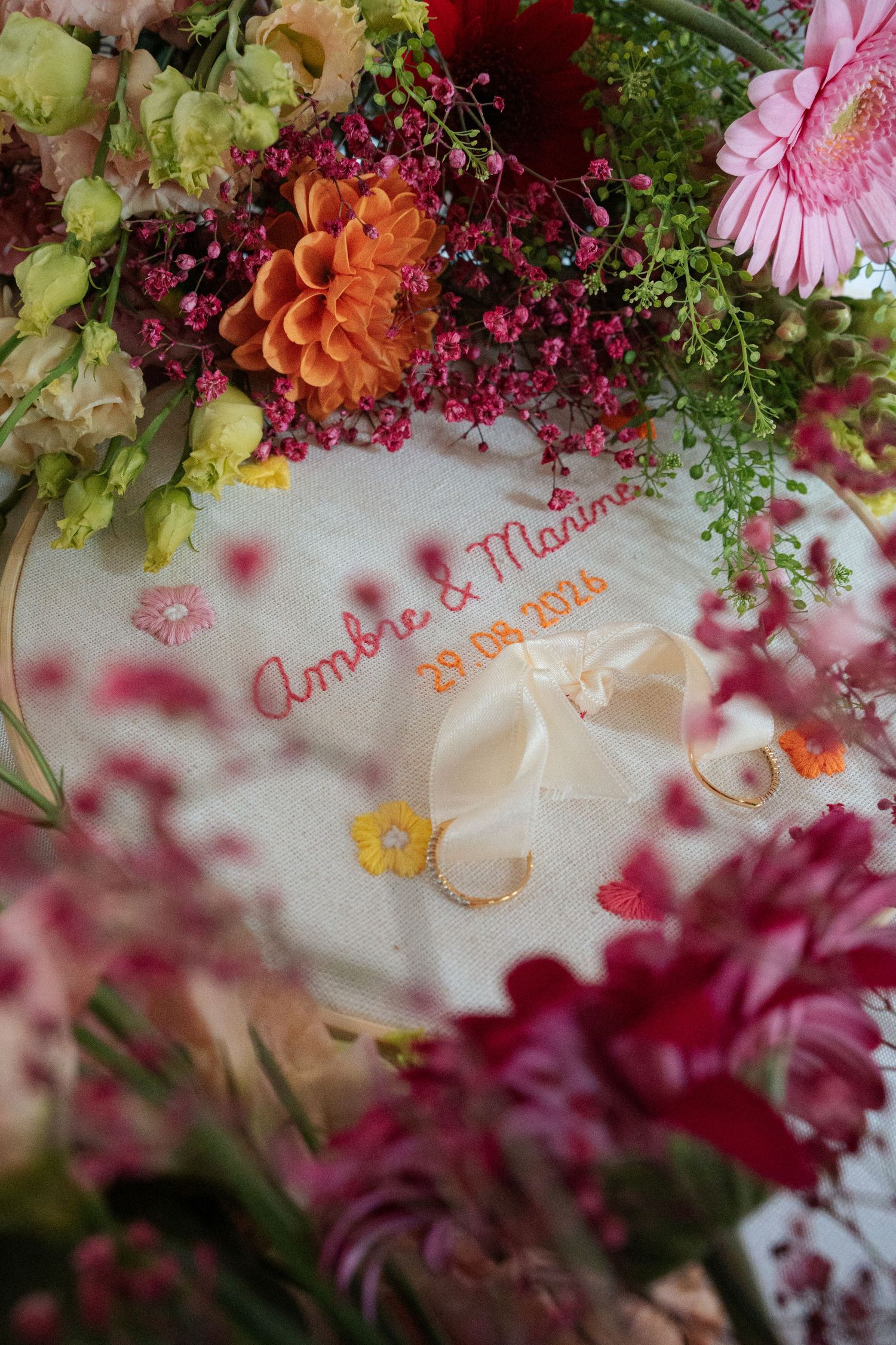 Embroidered wedding ring holder reading Ambre & Maxime 29.09.2024 surrounded by colorful pink and orange floral arrangements.
