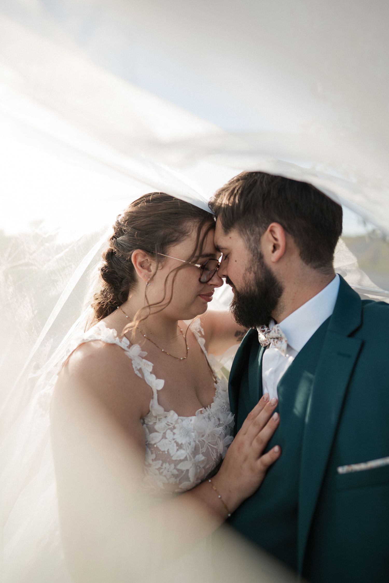 Bride and groom share an intimate moment under a flowing veil, with the groom in a teal suit and bride in lace dress.