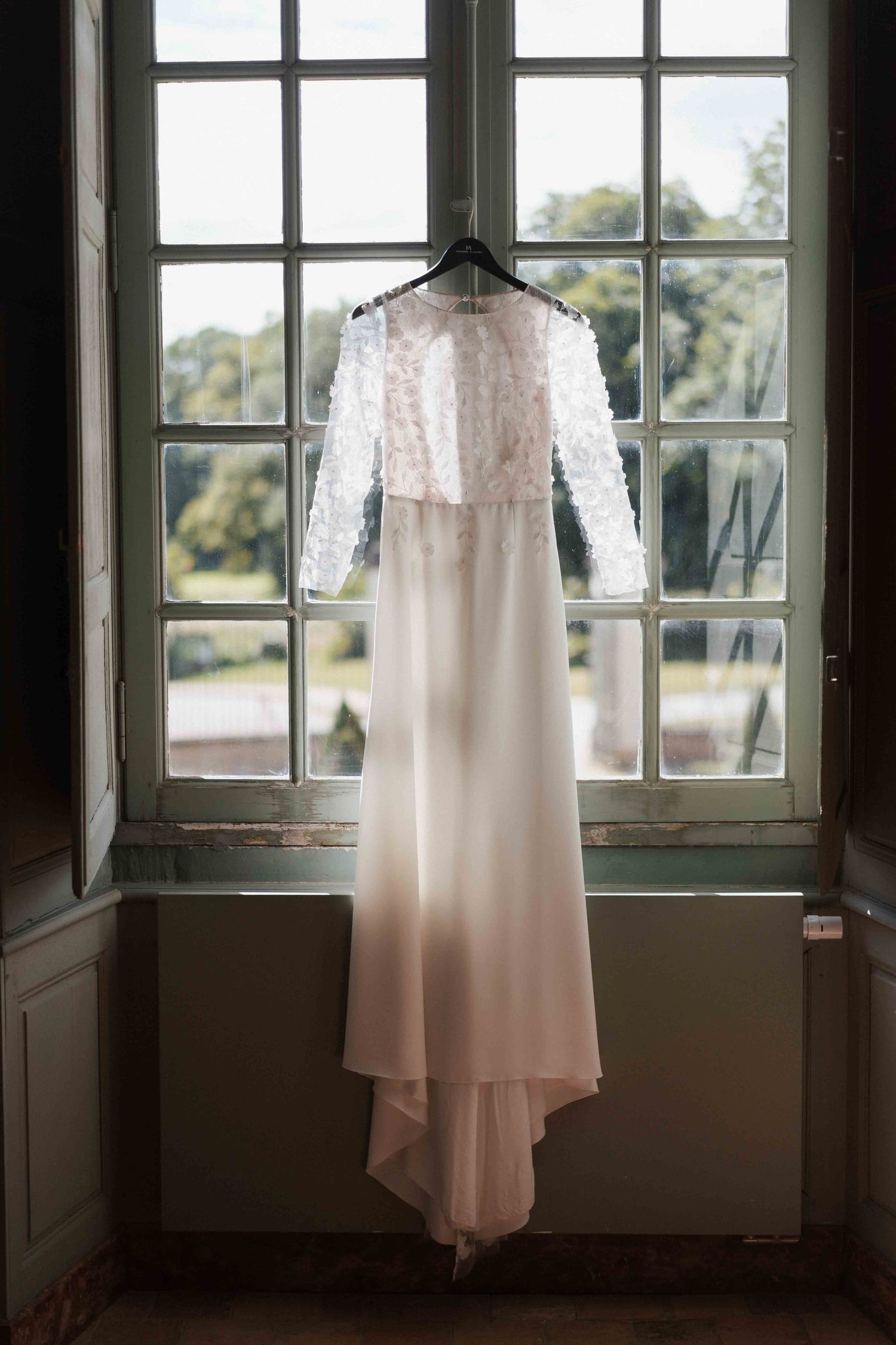 Élégante robe de mariée blanche à corsage en dentelle, suspendue à un cintre devant une fenêtre ancienne donnant sur un jardin.