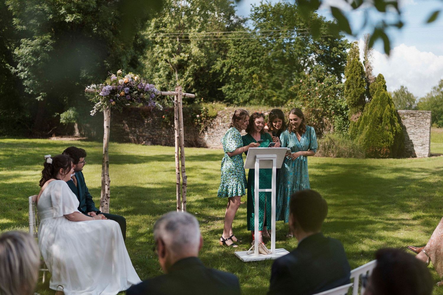 Discours lors d’une cérémonie laïque en plein air – Trois intervenantes lisent un texte devant les mariés, sous une arche fleurie.
