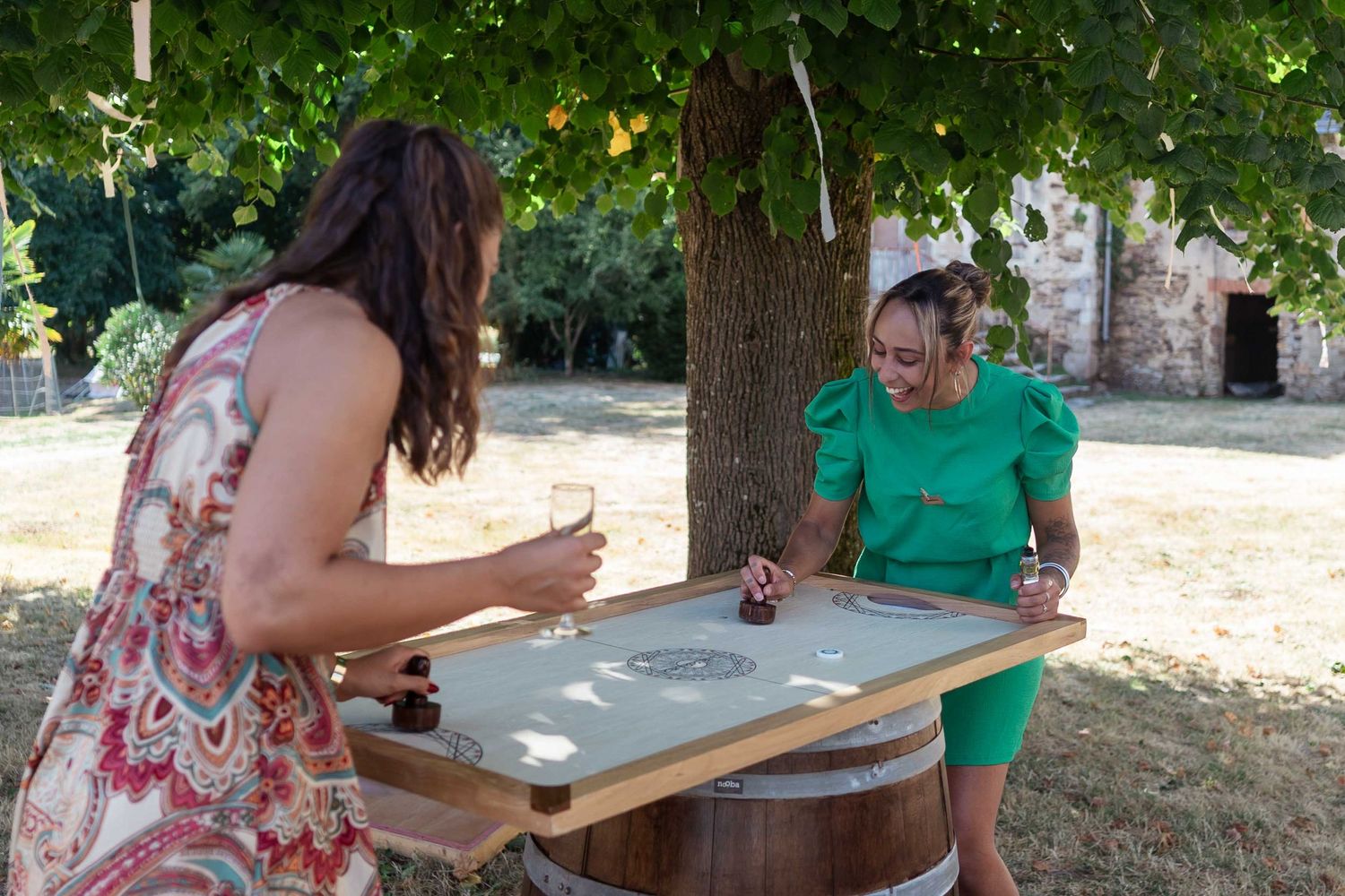 Deux femmes jouent à un jeu de société sur une table en forme de tonneau, à l'extérieur, sous un arbre ombragé.