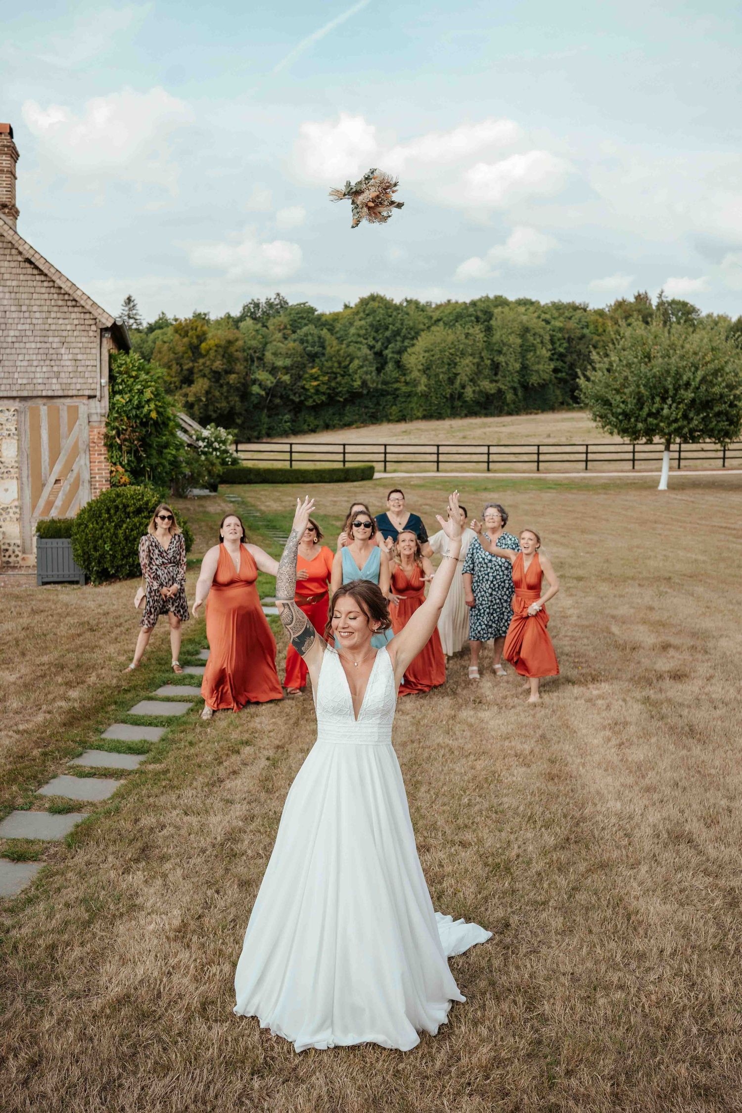 La mariée lance son bouquet de mariée à ses demoiselles d'honneur, ravies et vêtues de robes colorées, sur une pelouse ensoleillée.