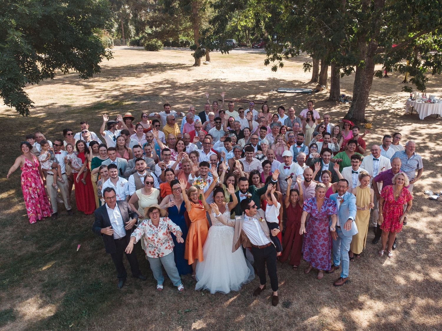 Aerial view of a large wedding party gathered in a heart shape under trees in an outdoor park setting.