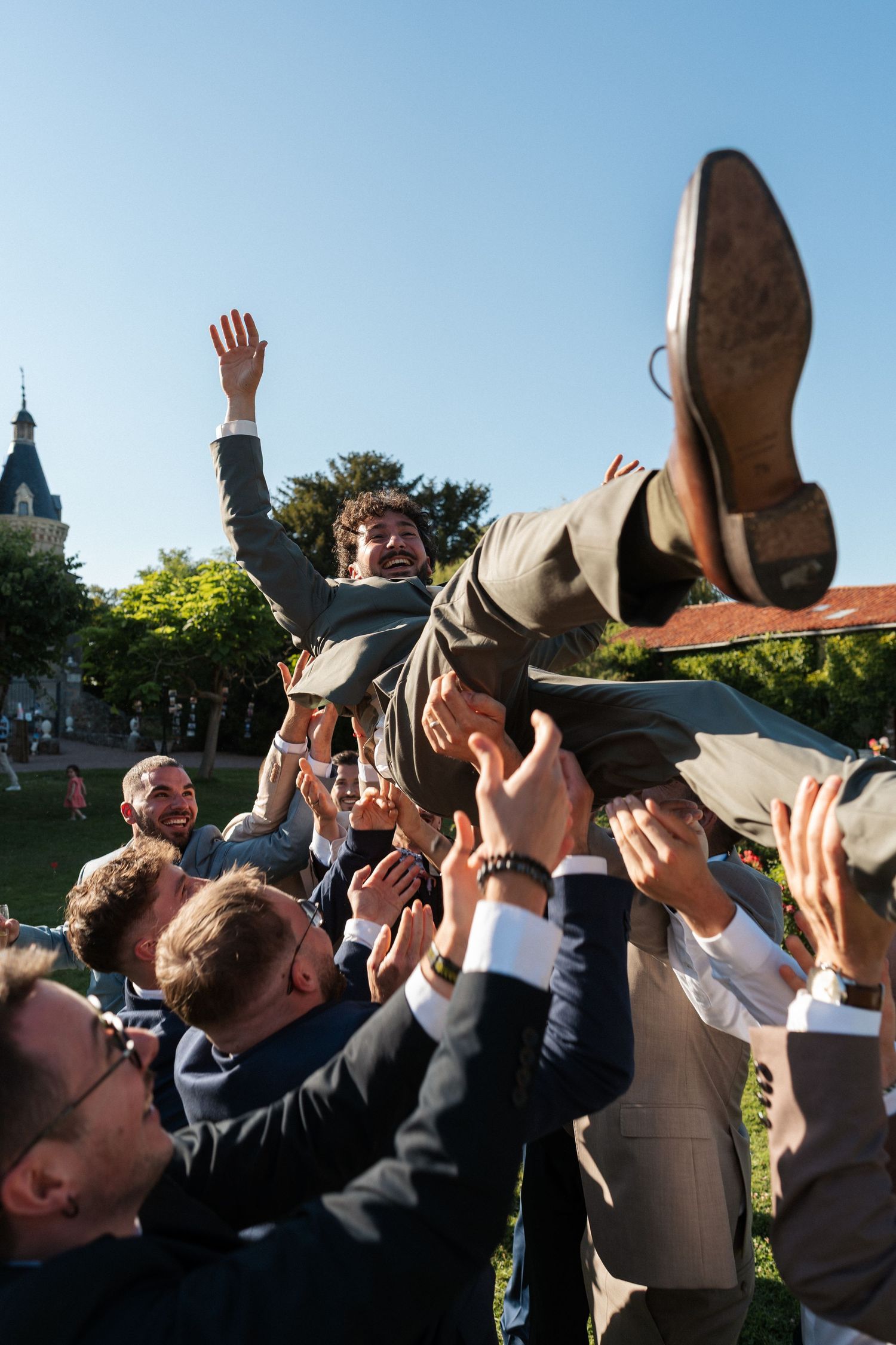 Men in suits tossing a groom into the air outdoors, celebrating at a wedding reception on a sunny day.