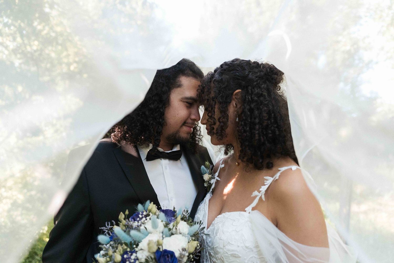 Les mariés partagent un moment intime sous le voile, avec un bouquet bleu en extérieur.