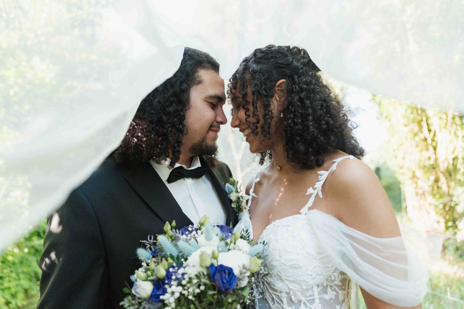 Les mariés partagent un moment romantique sous un voile flottant, la mariée tenant un bouquet bleu et blanc.