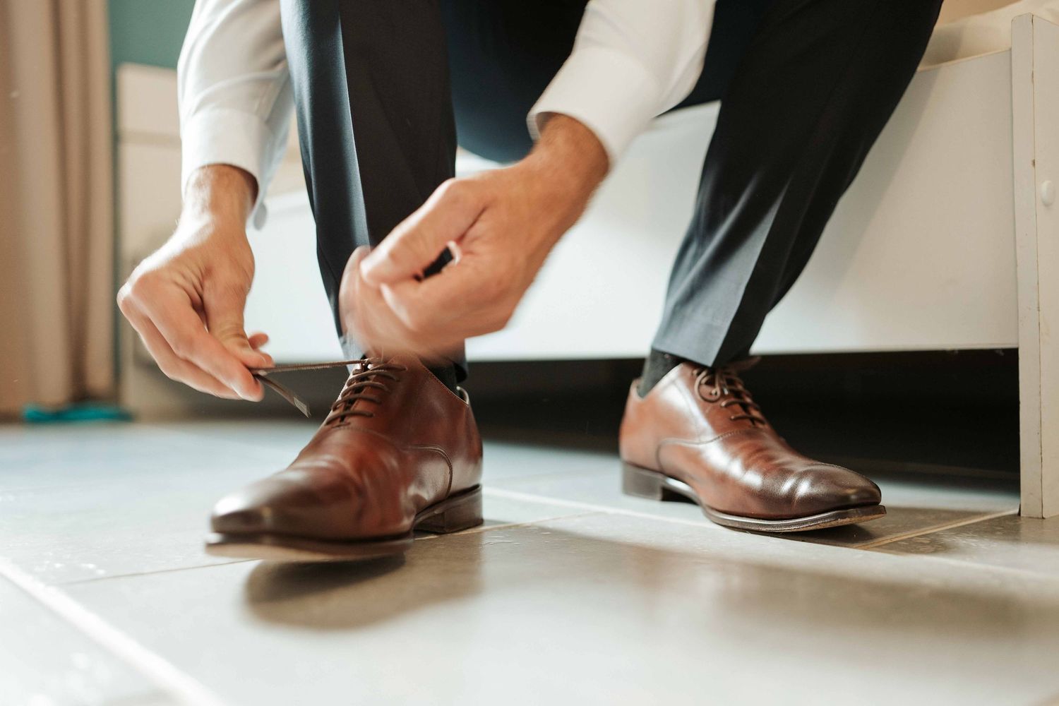 Un homme, assis, lace des chaussures Oxford en cuir marron, vêtu d'un pantalon noir et d'une chemise blanche.