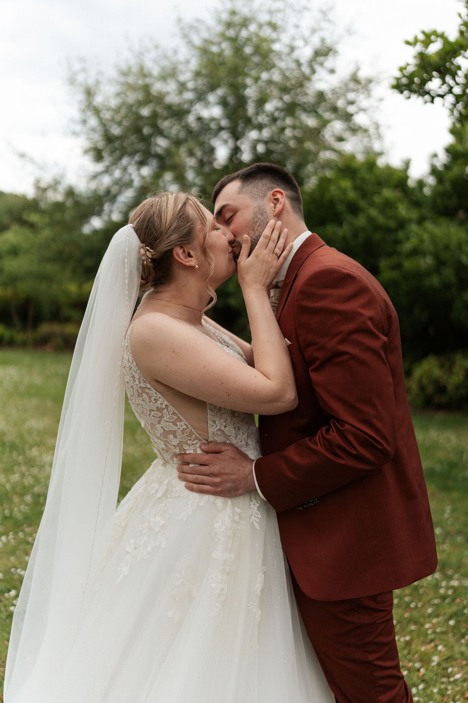 A bride and groom share a romantic kiss outdoors, she in a lace wedding gown with veil, he in a burgundy suit.