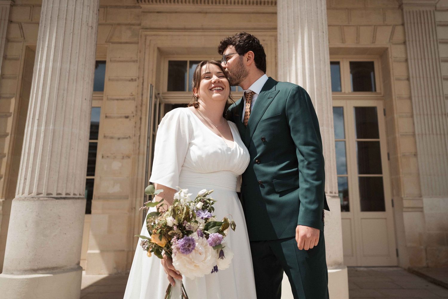 De jeunes mariés heureux s'enlacent devant un la mairie d'Angers la mariée tenant un bouquet de fleurs sauvages.