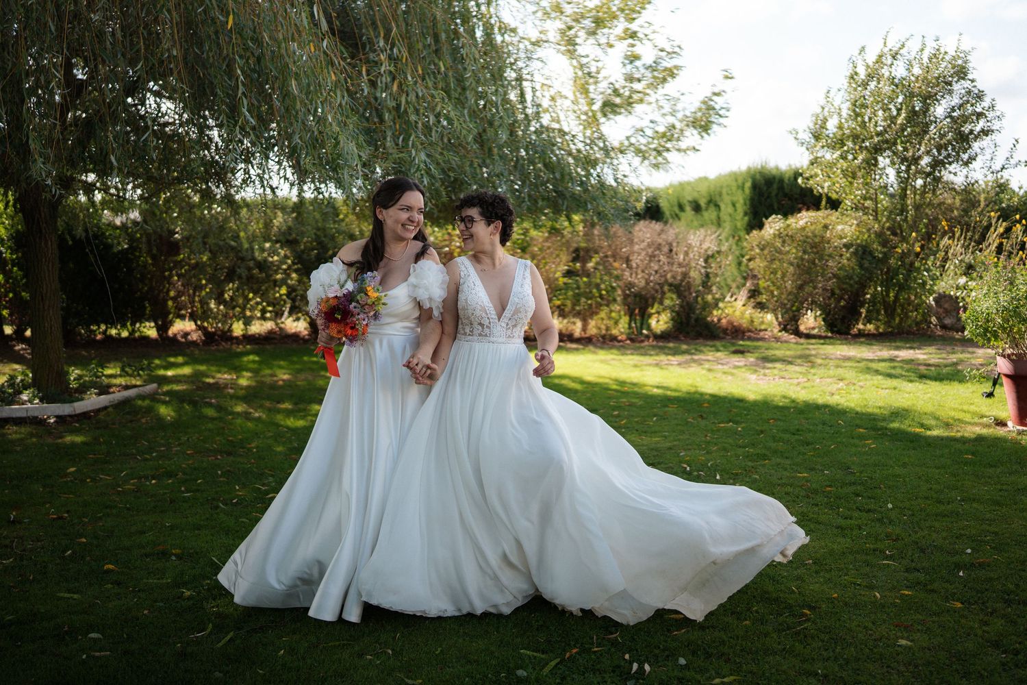 Two brides in white wedding gowns smiling together outdoors in a lush green garden on their wedding day.