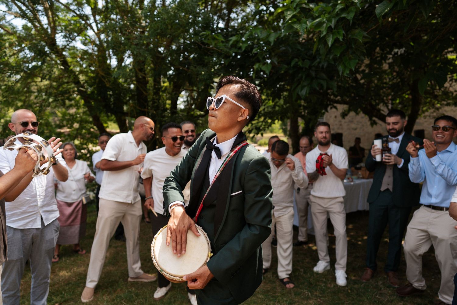 Person in green suit and white sunglasses playing tambourine outdoors at a celebration with guests clapping.
