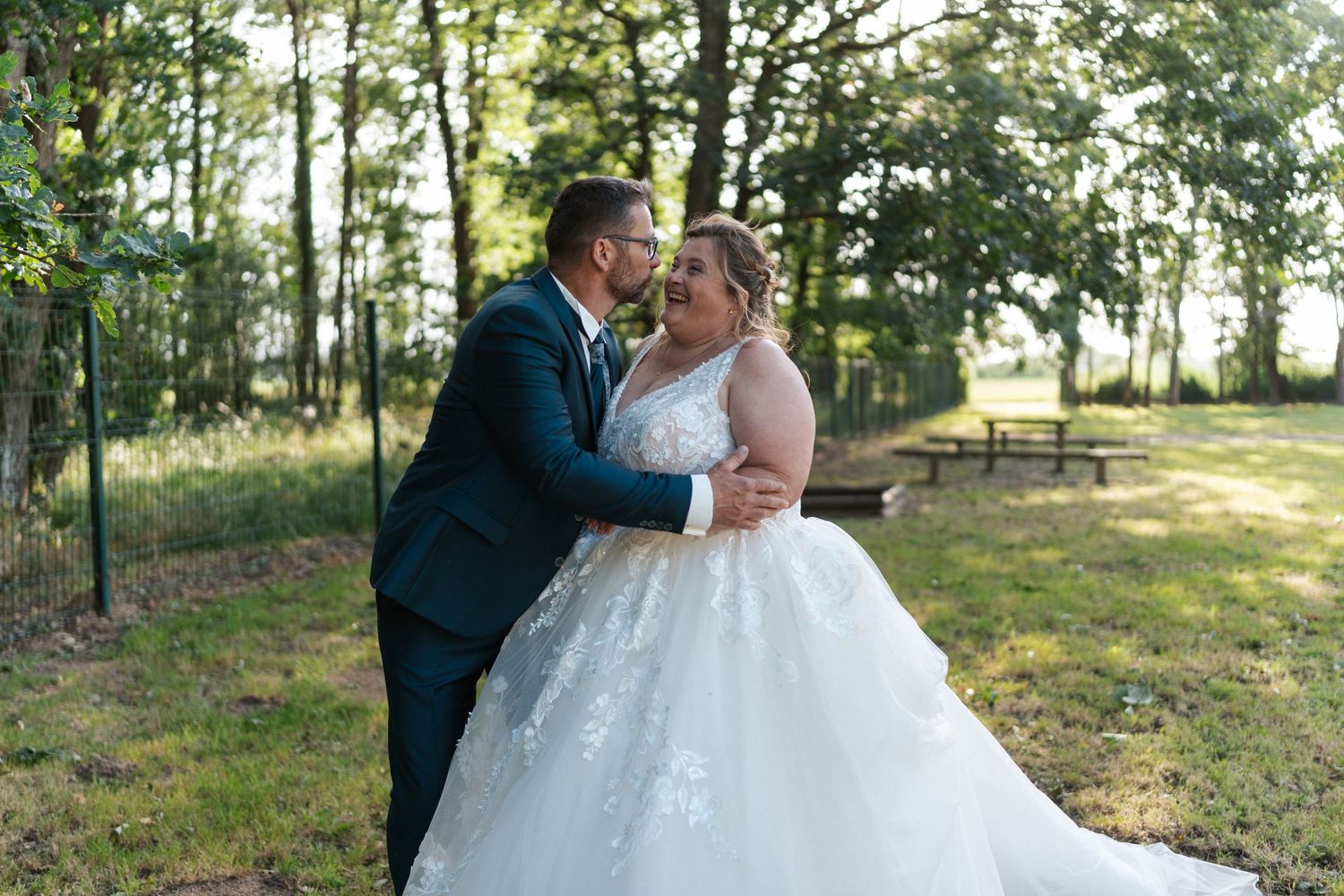 Bride in lace ball gown and groom in navy suit share a romantic moment outdoors near trees at a wedding.