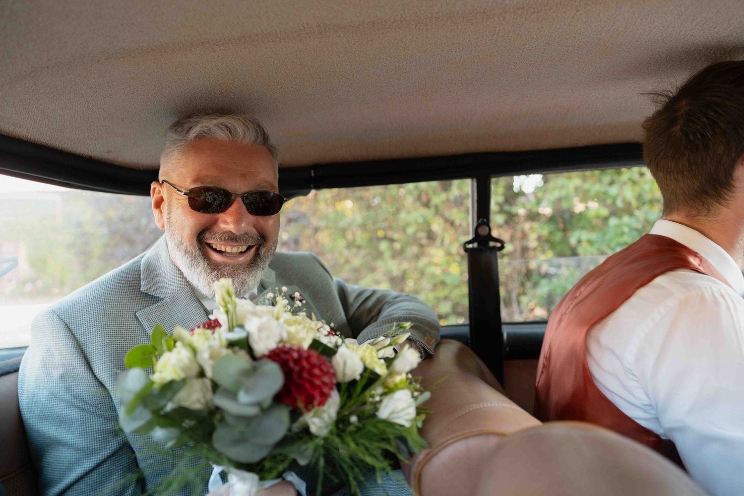 Un homme souriant, portant des lunettes de soleil et tenant un bouquet de mariée, à l'intérieur d'une voiture ancienne de collection.