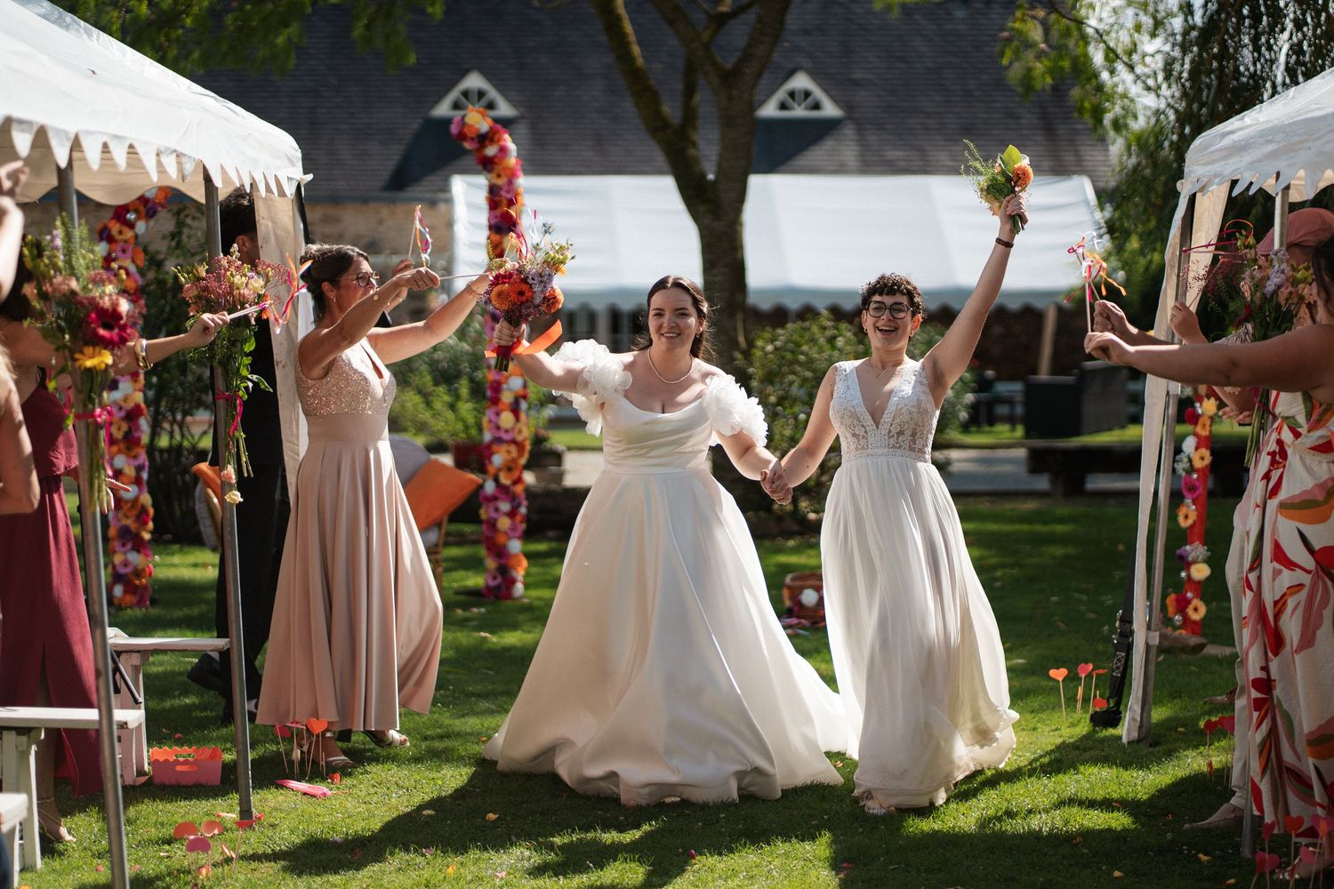 Two brides in white gowns celebrate with bridesmaids at an outdoor fall wedding ceremony.