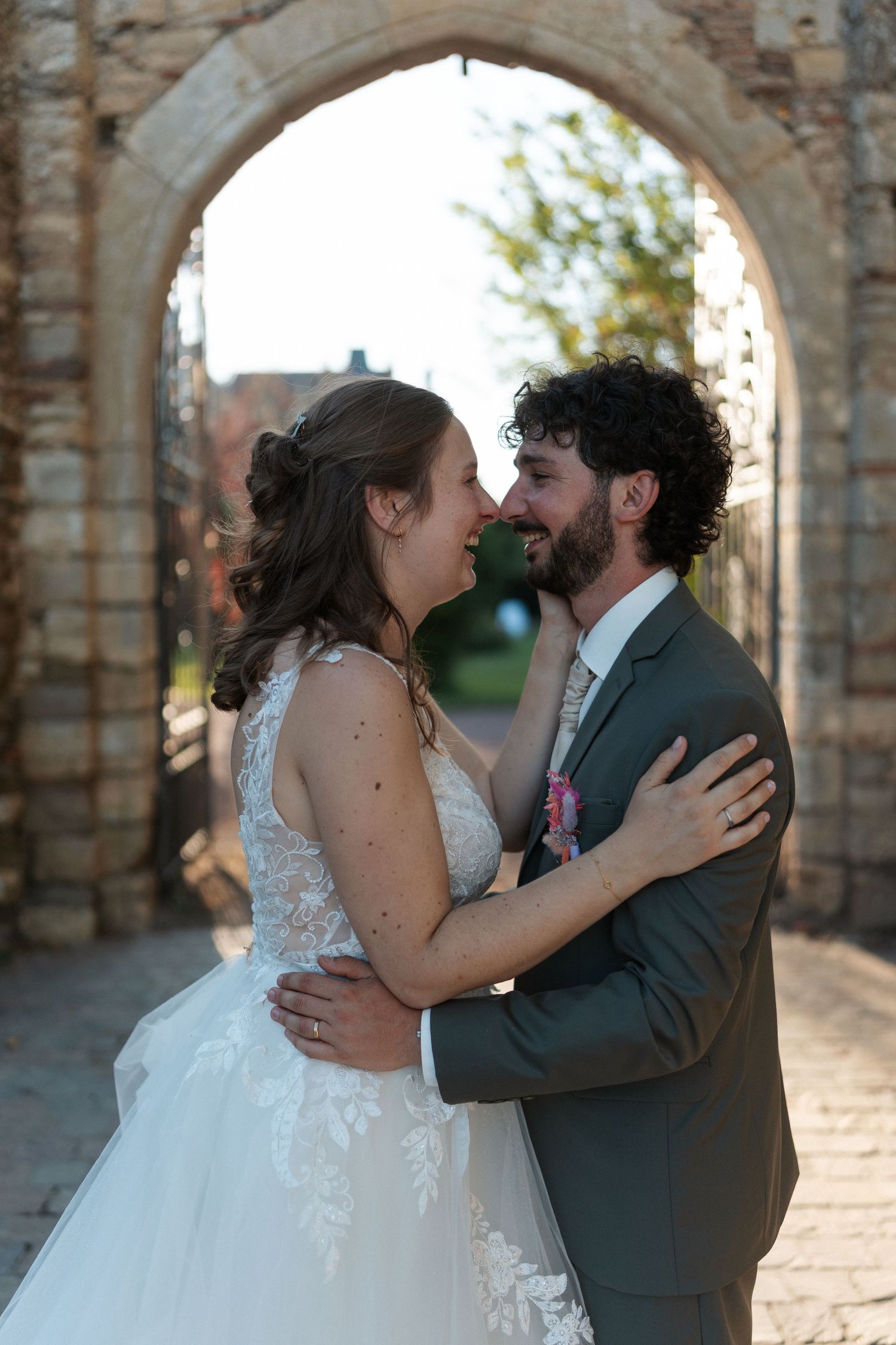 Bride and groom embracing and smiling at each other under a stone arch in a romantic wedding portrait.