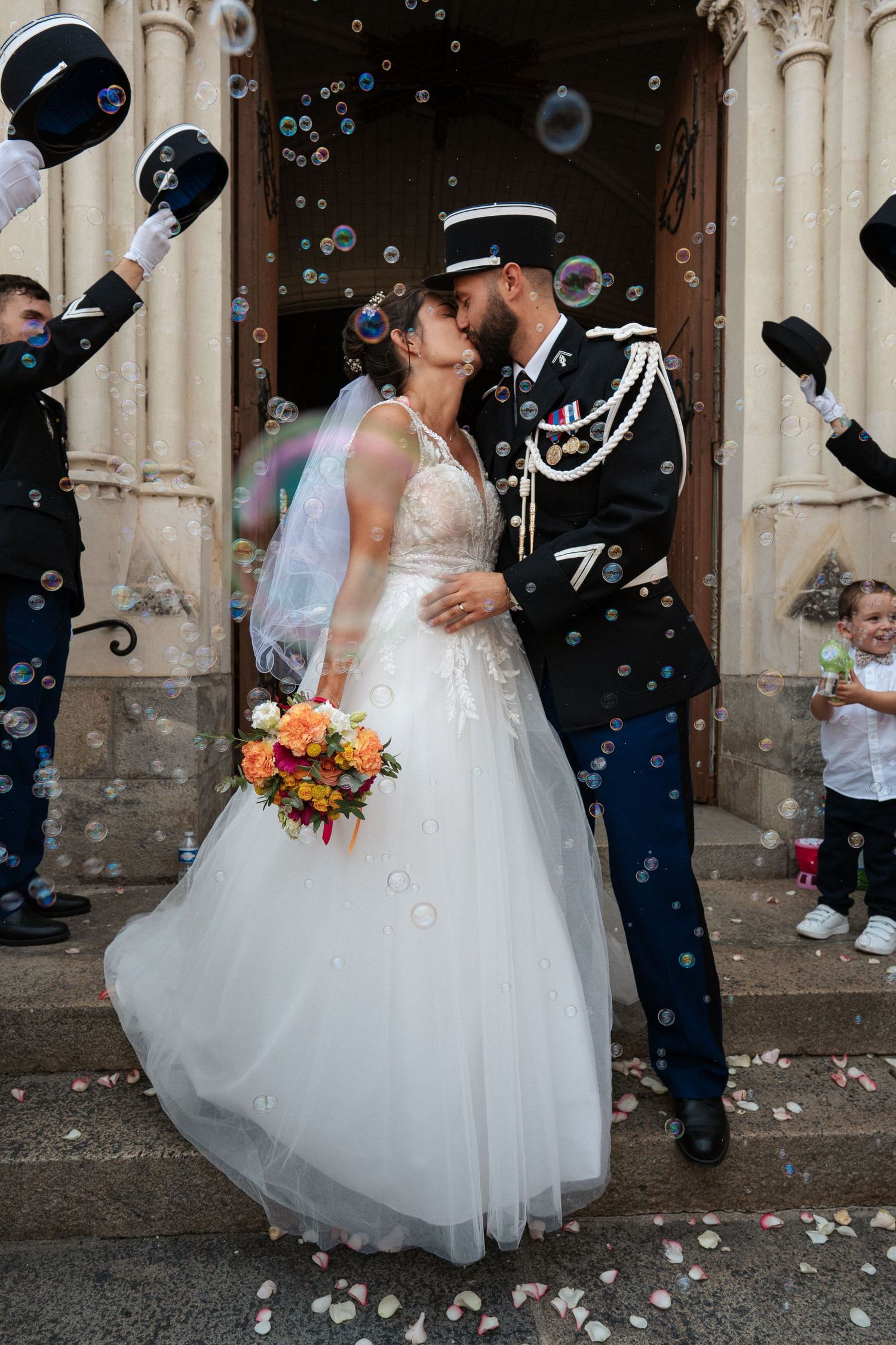 Newlywed couple kissing outside church surrounded by soap bubbles, bride in white gown holding bouquet, groom in military uniform.