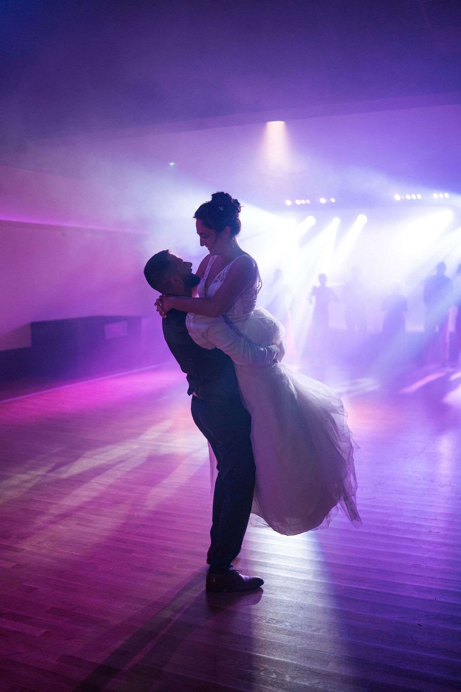 Groom lifting bride in wedding dress during romantic first dance on purple-lit dance floor.