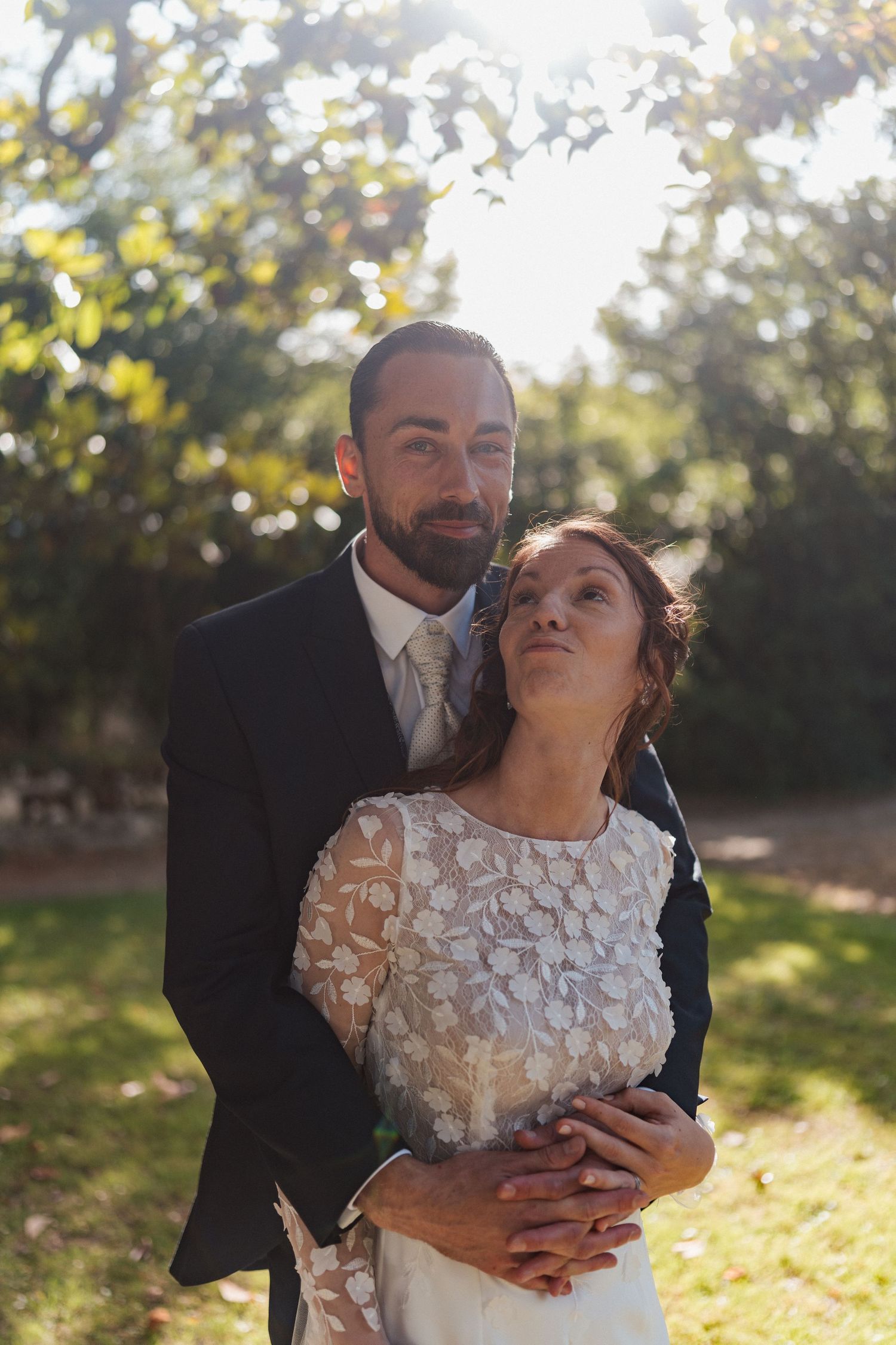 Dans un jardin ensoleillé, un marié enlace sa femme par derrière, tous deux souriant doucement dans leurs élégantes tenues de mariage.