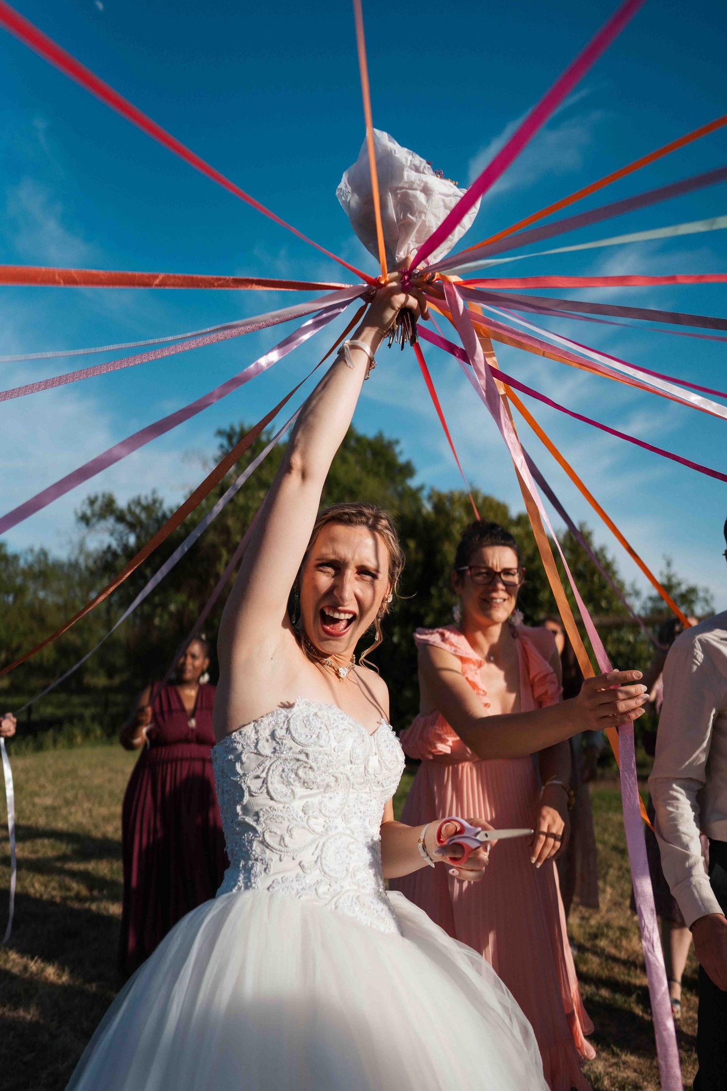 Une mariée rayonnante en robe blanche tient des rubans colorés au-dessus de sa tête lors de la traditionnelle cérémonie de lancer de bouquet